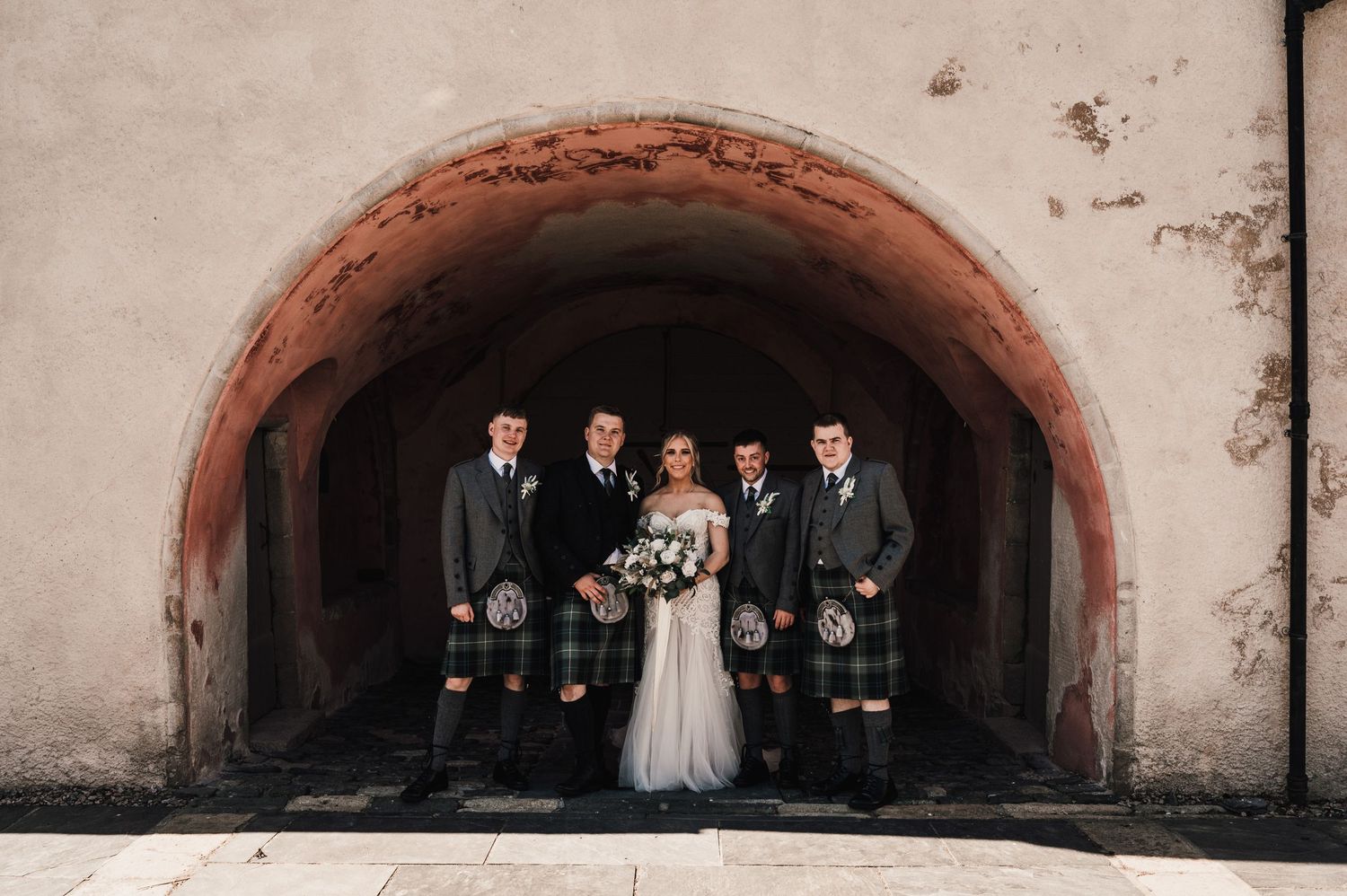 Wedding party in kilts and formal attire posed under stone archway.