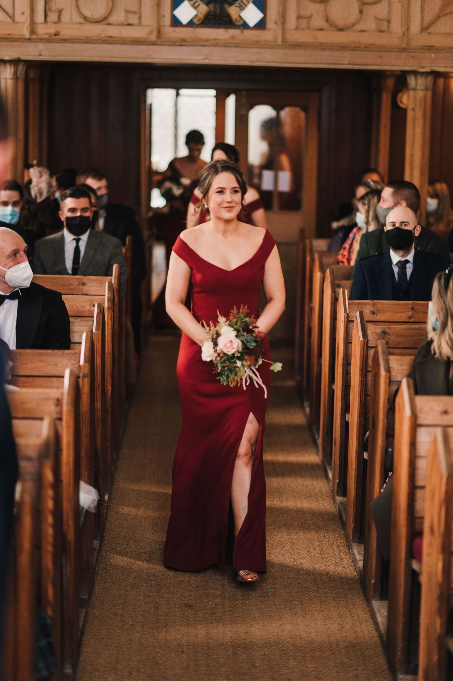 A bridesmaid walks down wooden church aisle in flowing burgundy gown with bouquet.