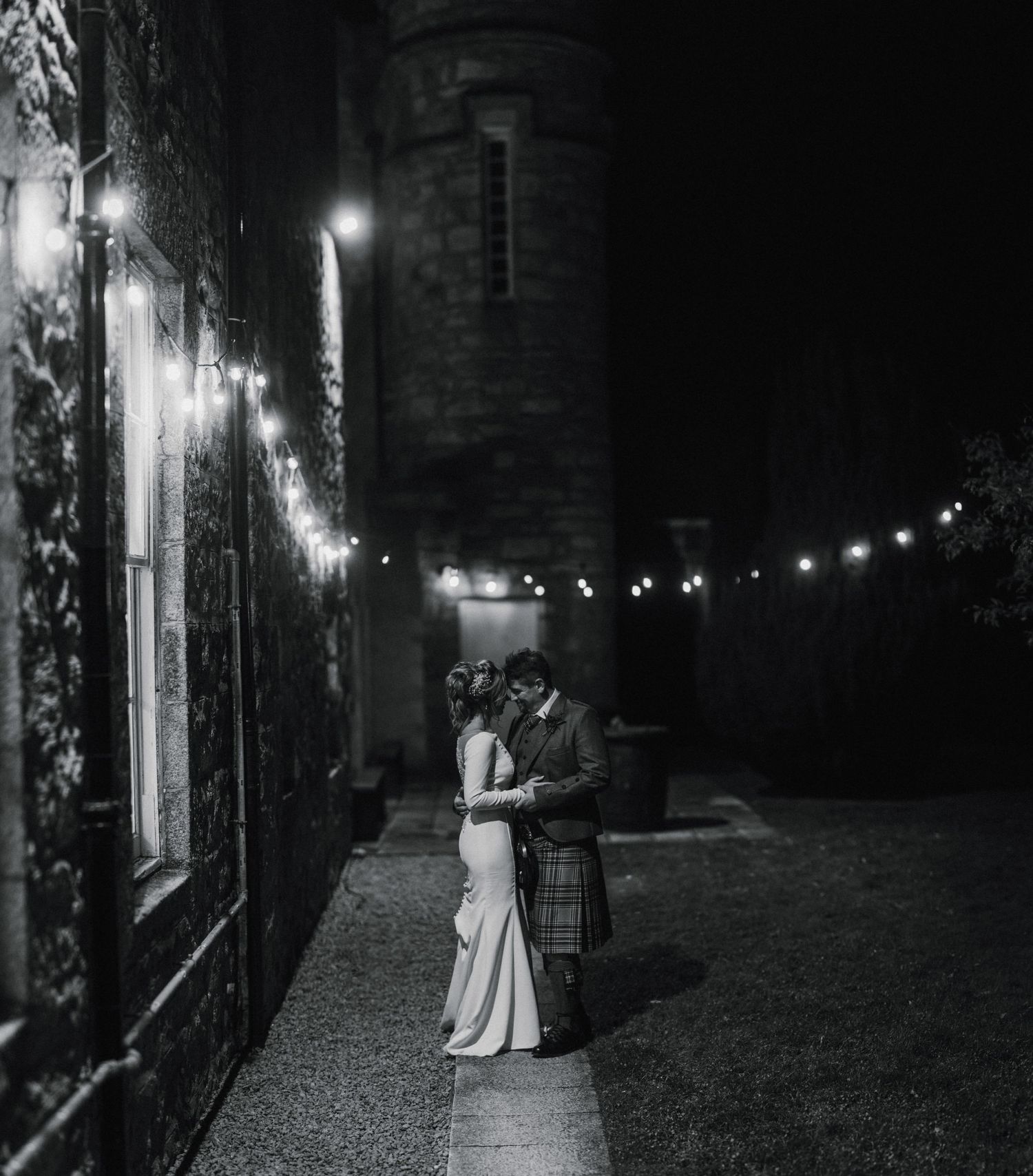 A romantic nighttime portrait of newlyweds under string lights along a stone building wall.