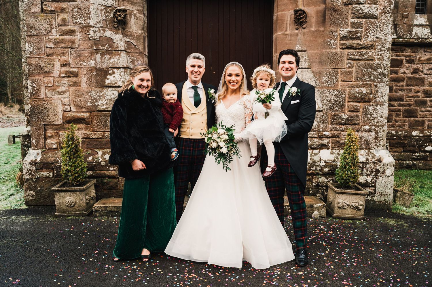 Wedding party members in elegant attire pose together outside historic church entrance.
