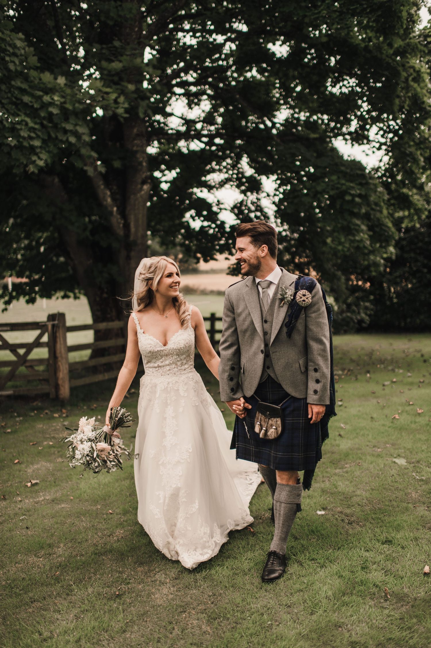 A couple in wedding attire walks together on grass, with the groom wearing a traditional Scottish kilt.