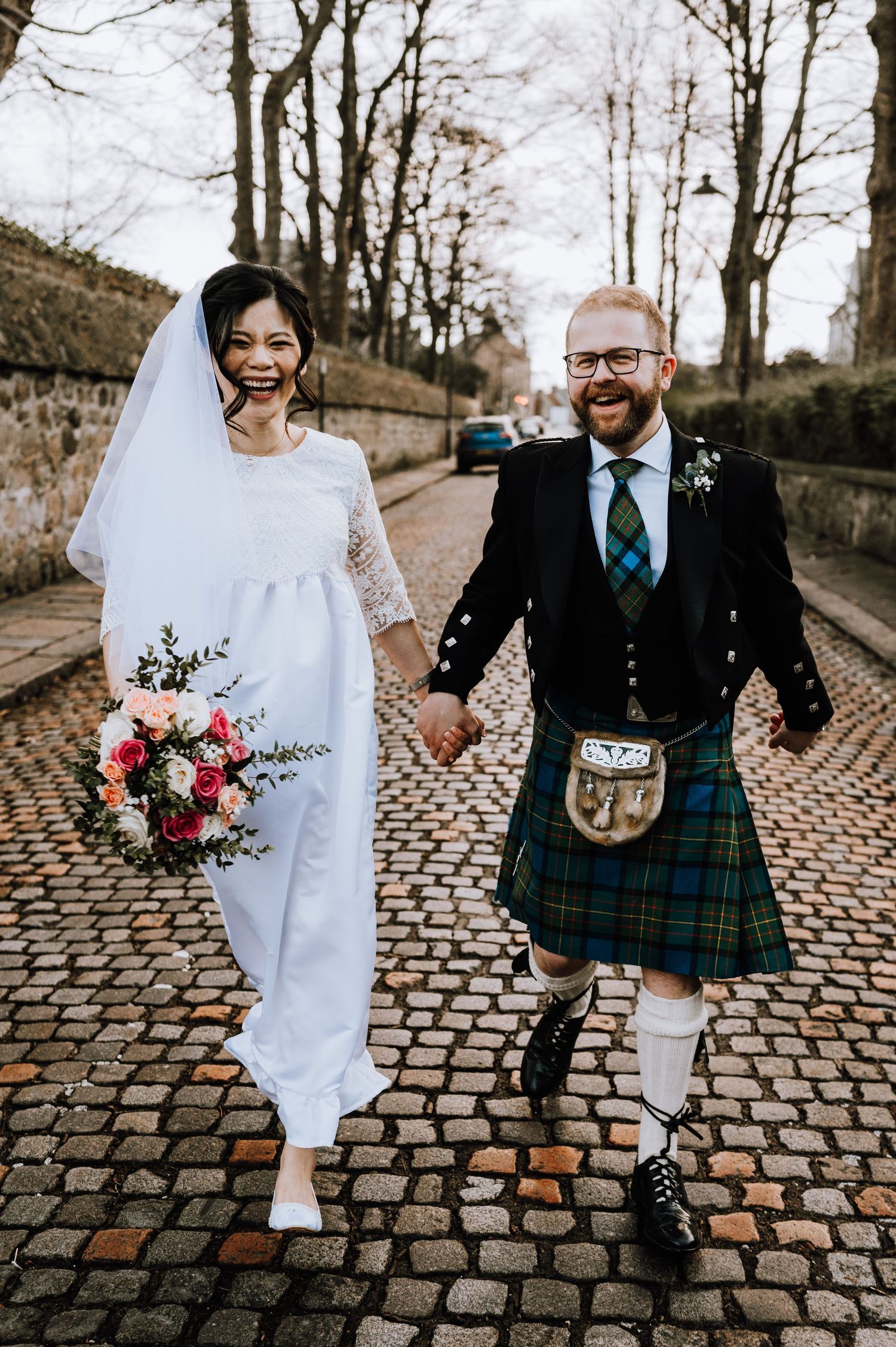 A joyful wedding couple walks hand in hand on cobblestones, with bride holding a colorful bouquet.