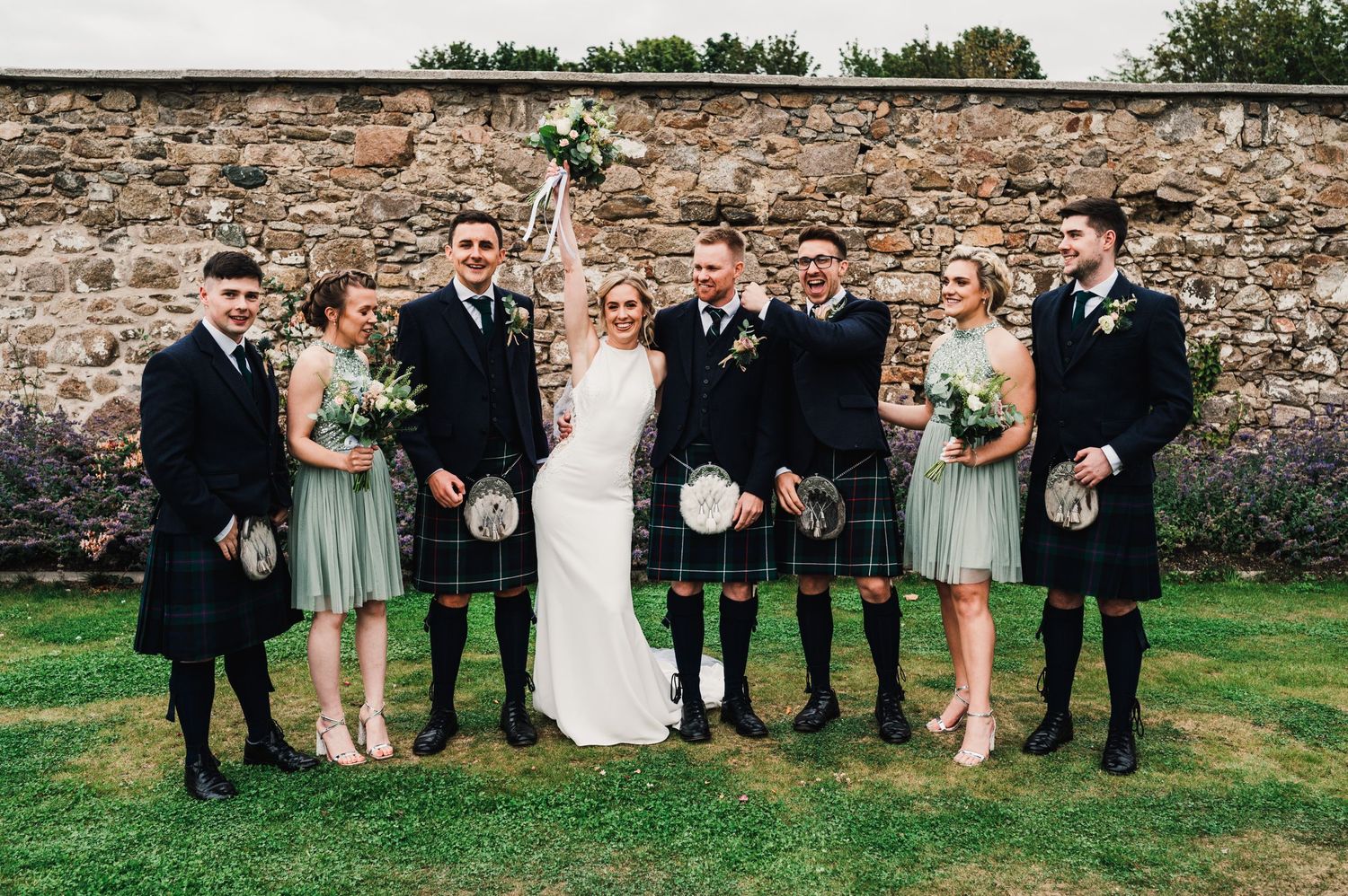 Wedding group in sage green dresses and traditional Scottish kilts stand together on lawn by stone wall.