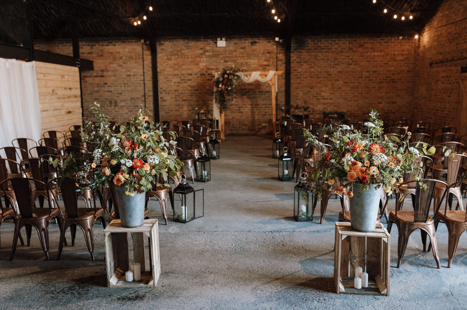Rustic wedding venue with wooden chairs and floral arrangements lining the aisle in an industrial brick space.