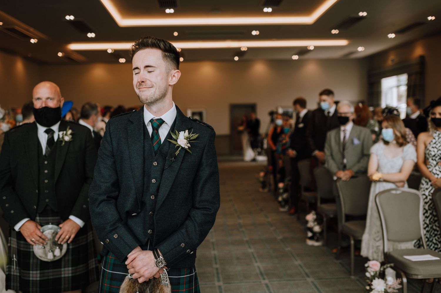 Attendee in dark suit and kilt stands at front of ceremony venue with guests seated.