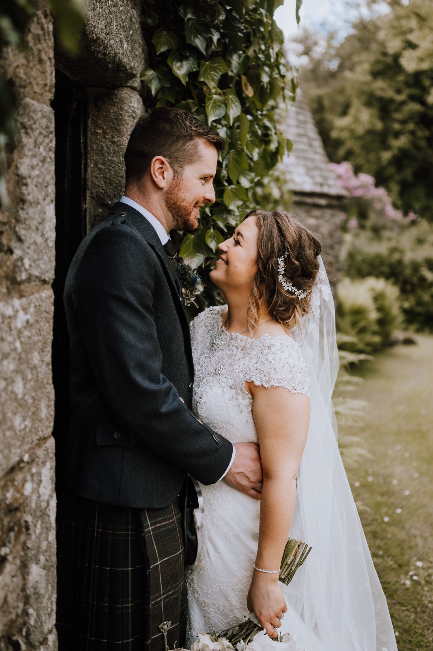 Wedding couple embraces by a stone wall with ivy, bride in lace and groom in traditional Scottish attire.
