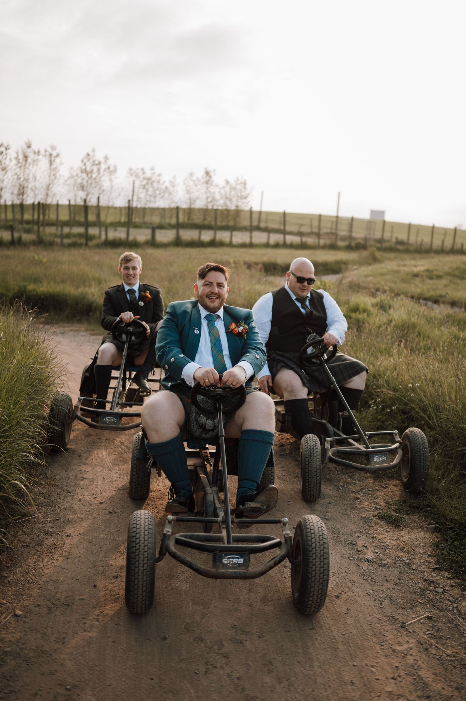 Wedding party rides go-karts on dirt path wearing traditional Scottish attire.