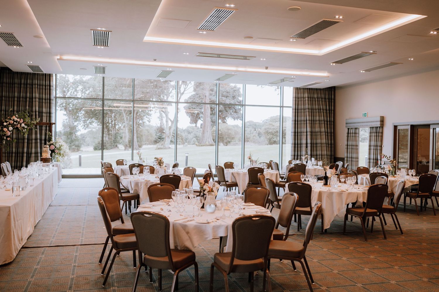 Modern wedding reception venue with round tables, white linens, and large windows overlooking natural scenery.