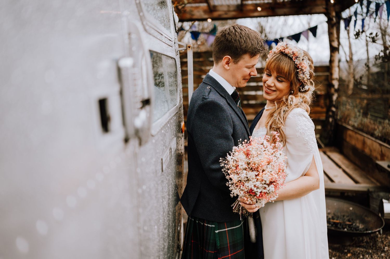 Scottish wedding couple embraces near white wall, bride holding delicate pink bouquet.