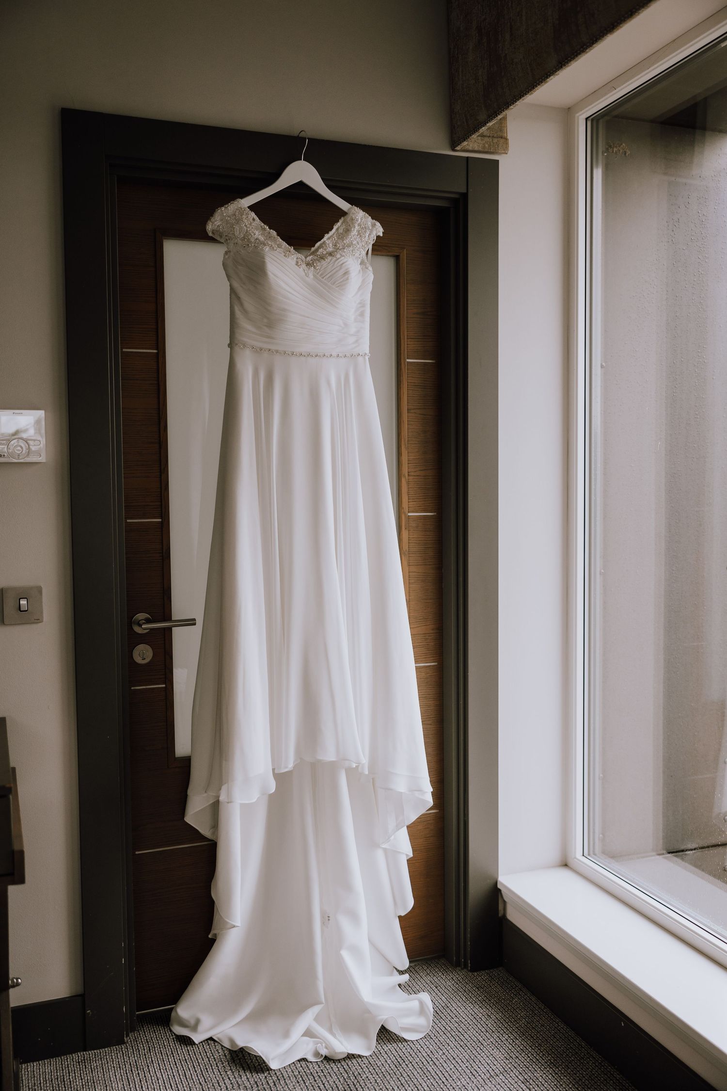 An elegant white wedding dress hangs on a dark wooden door in a hotel room.