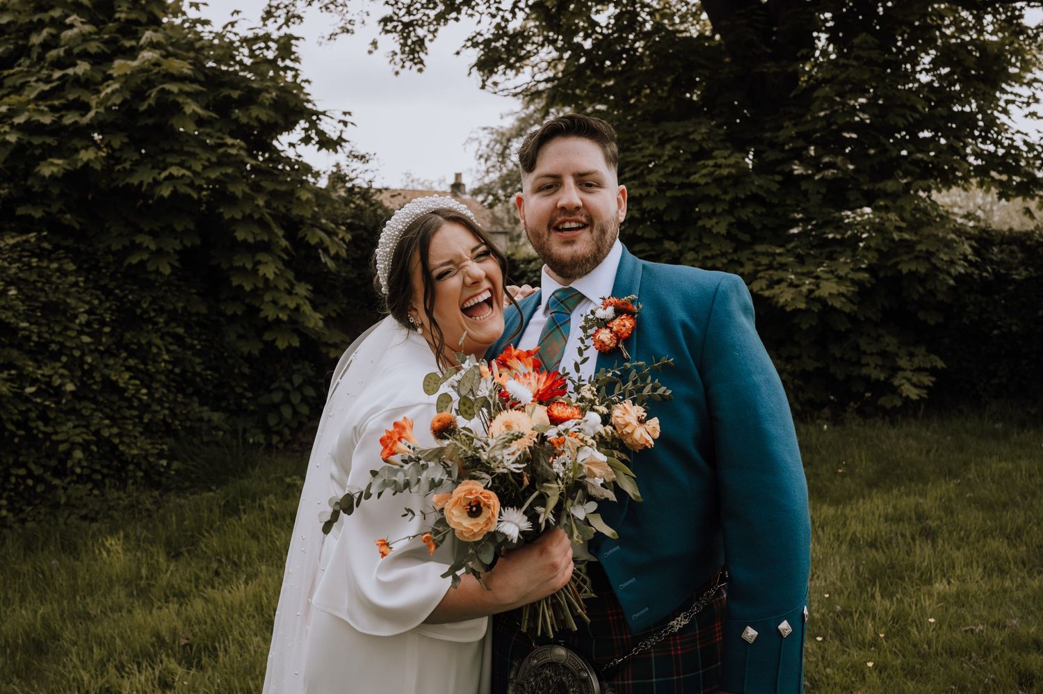 Joyful newlyweds share a laugh while holding autumn-colored bouquet outdoors.