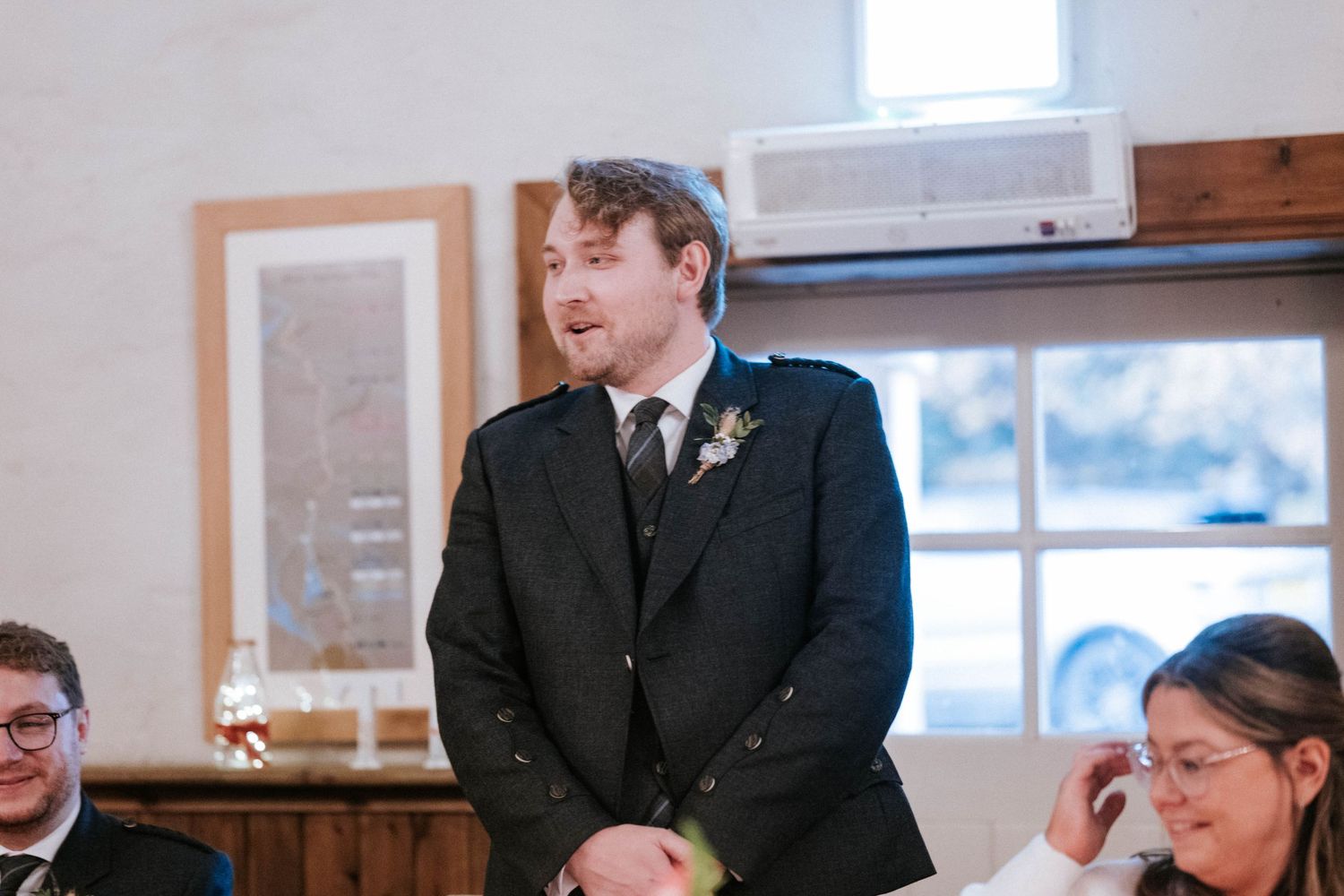 Wedding party members gather around tables during an indoor reception event.