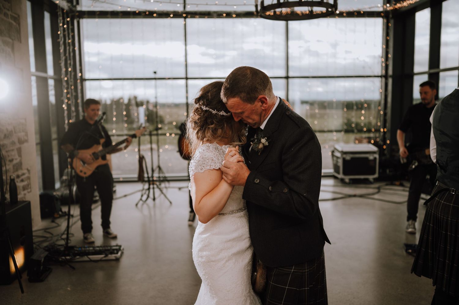 Intimate wedding dance moment in a venue with floor-to-ceiling windows and live band in background.