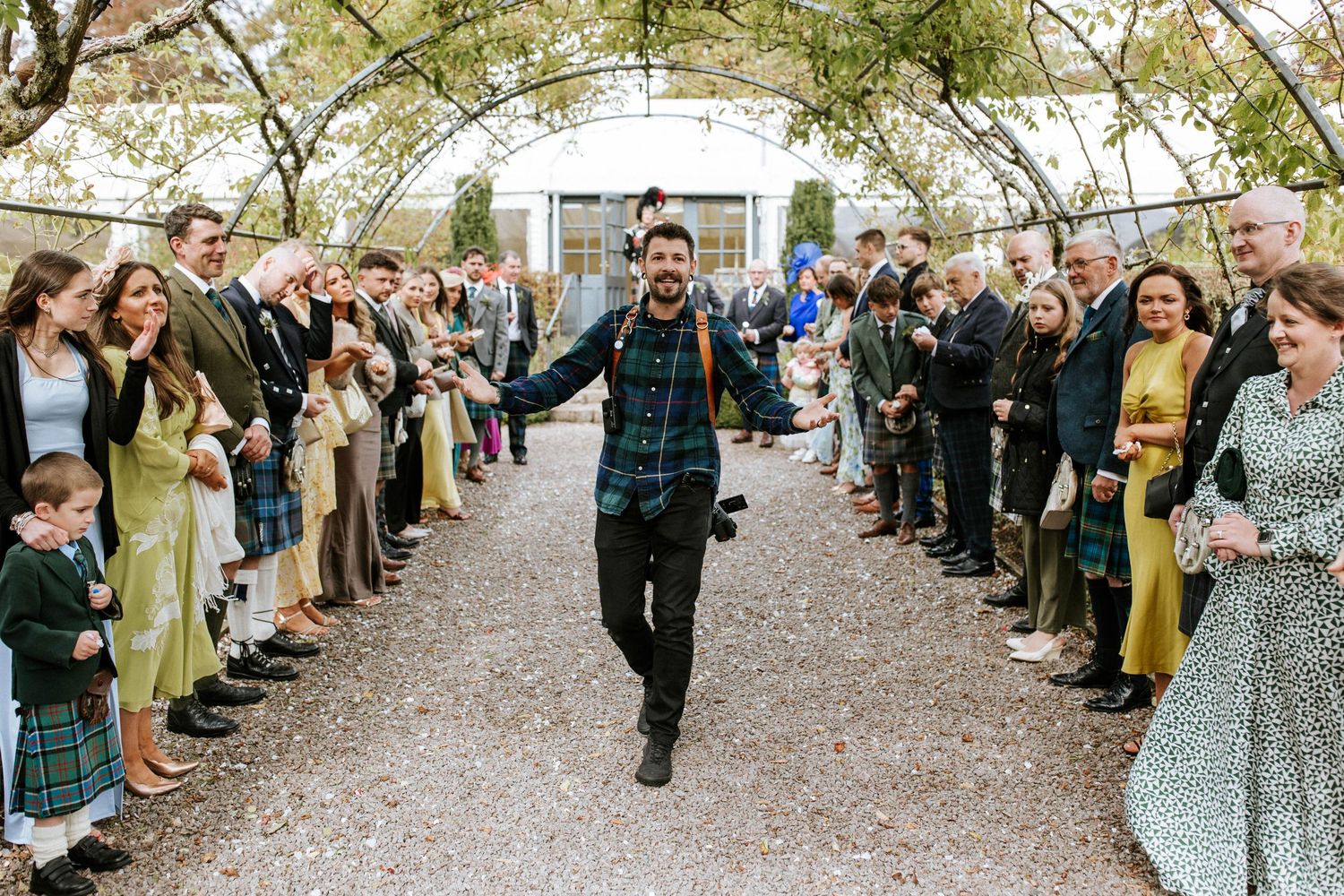 Wedding guests form a celebratory walkway under trees during an outdoor ceremony.