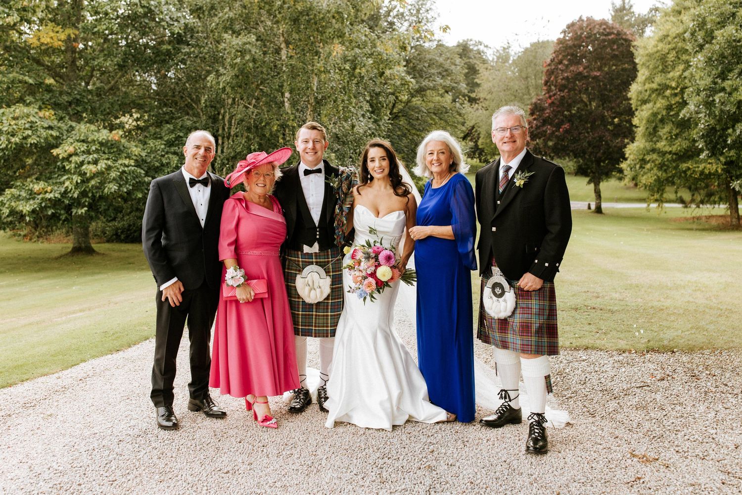 Wedding party portrait outdoors with Scottish kilts and formal attire against garden backdrop.