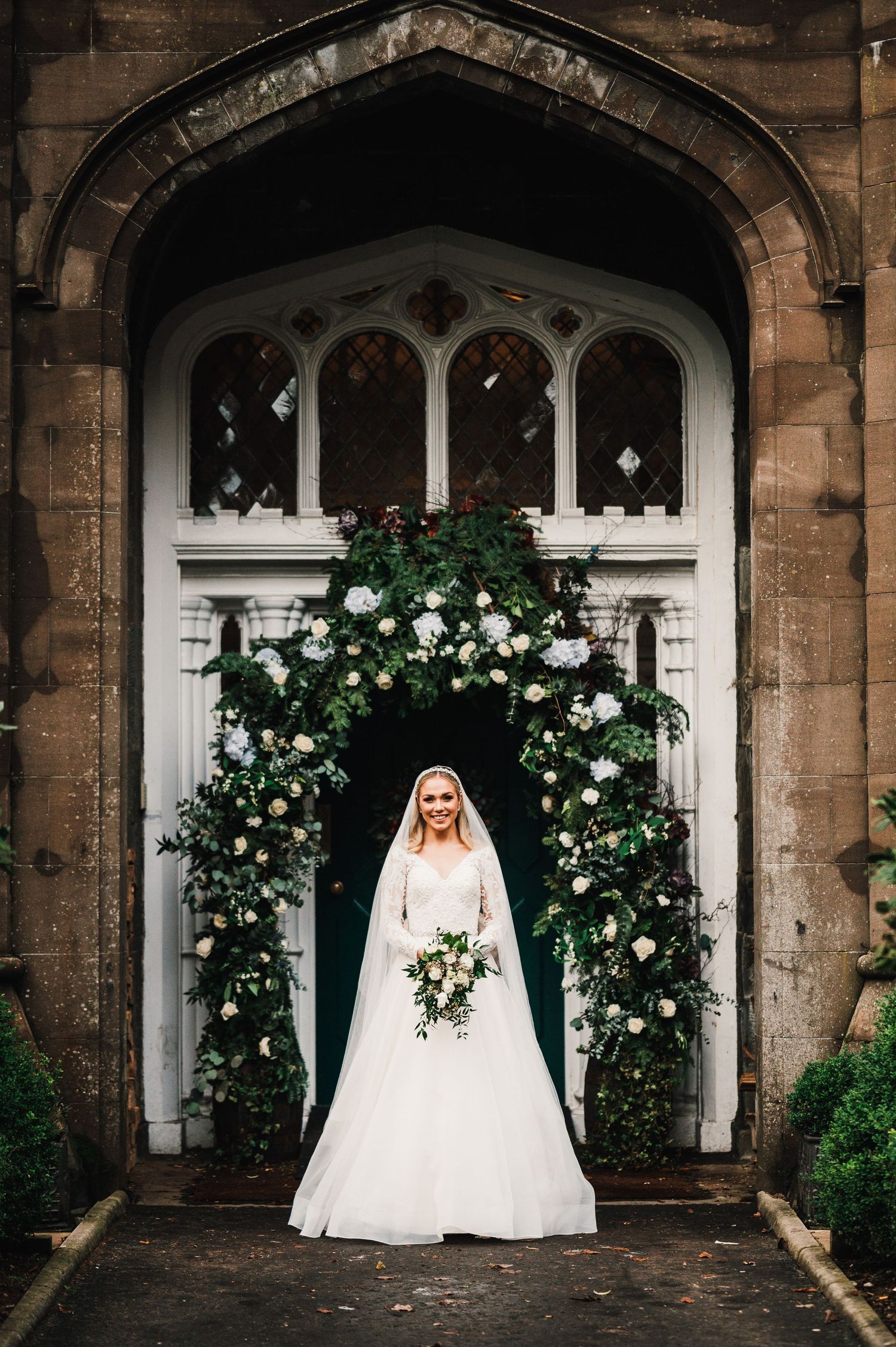 Aberdeen Wedding Photography. Bride in white gown poses under gothic stone archway decorated with climbing white roses.