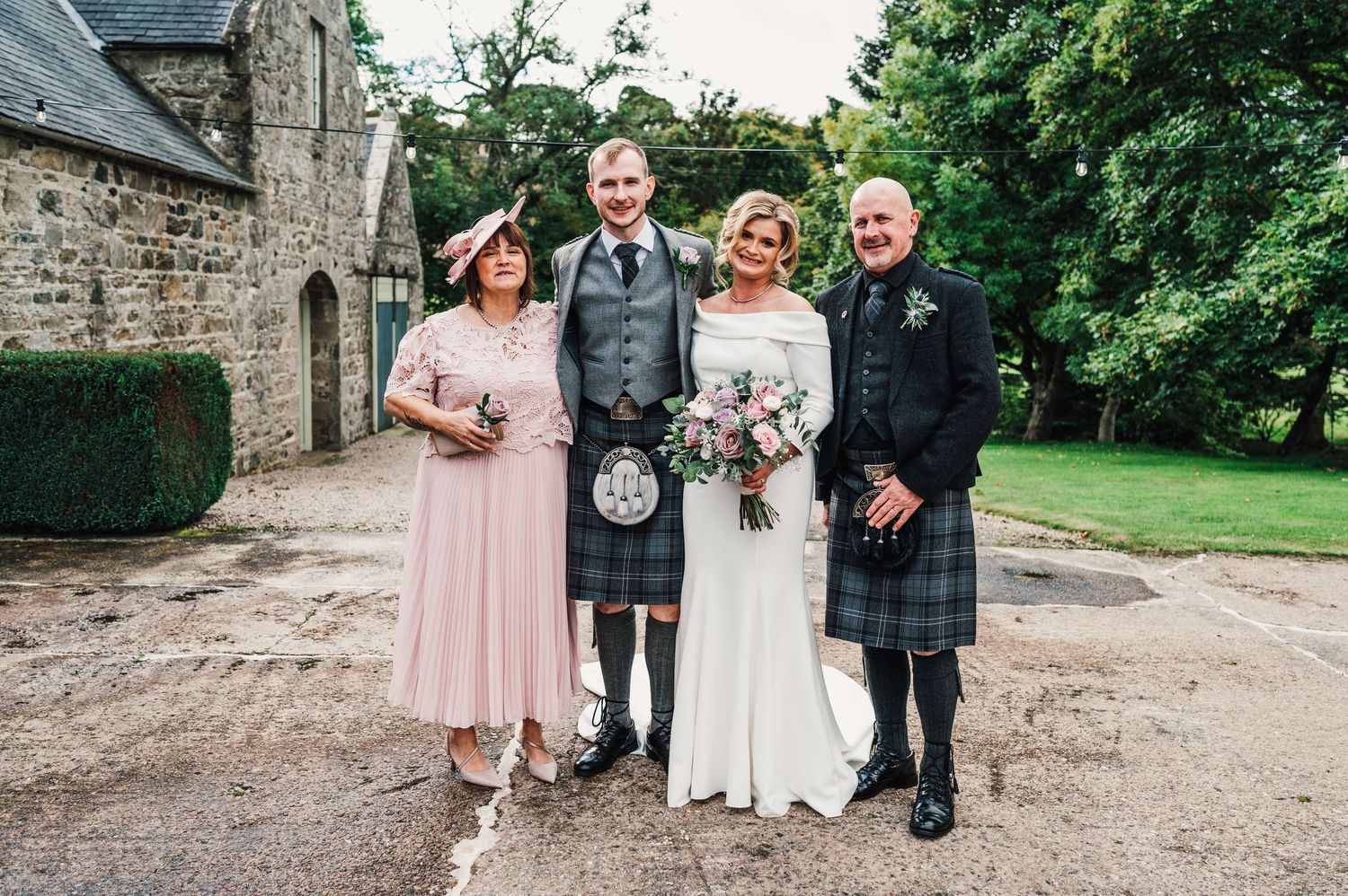 Wedding party poses for group photos outside a rustic stone building surrounded by greenery.