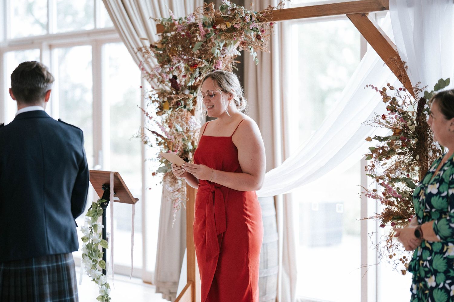 Wedding ceremony setup with dried floral arrangements and draped curtains by large windows.