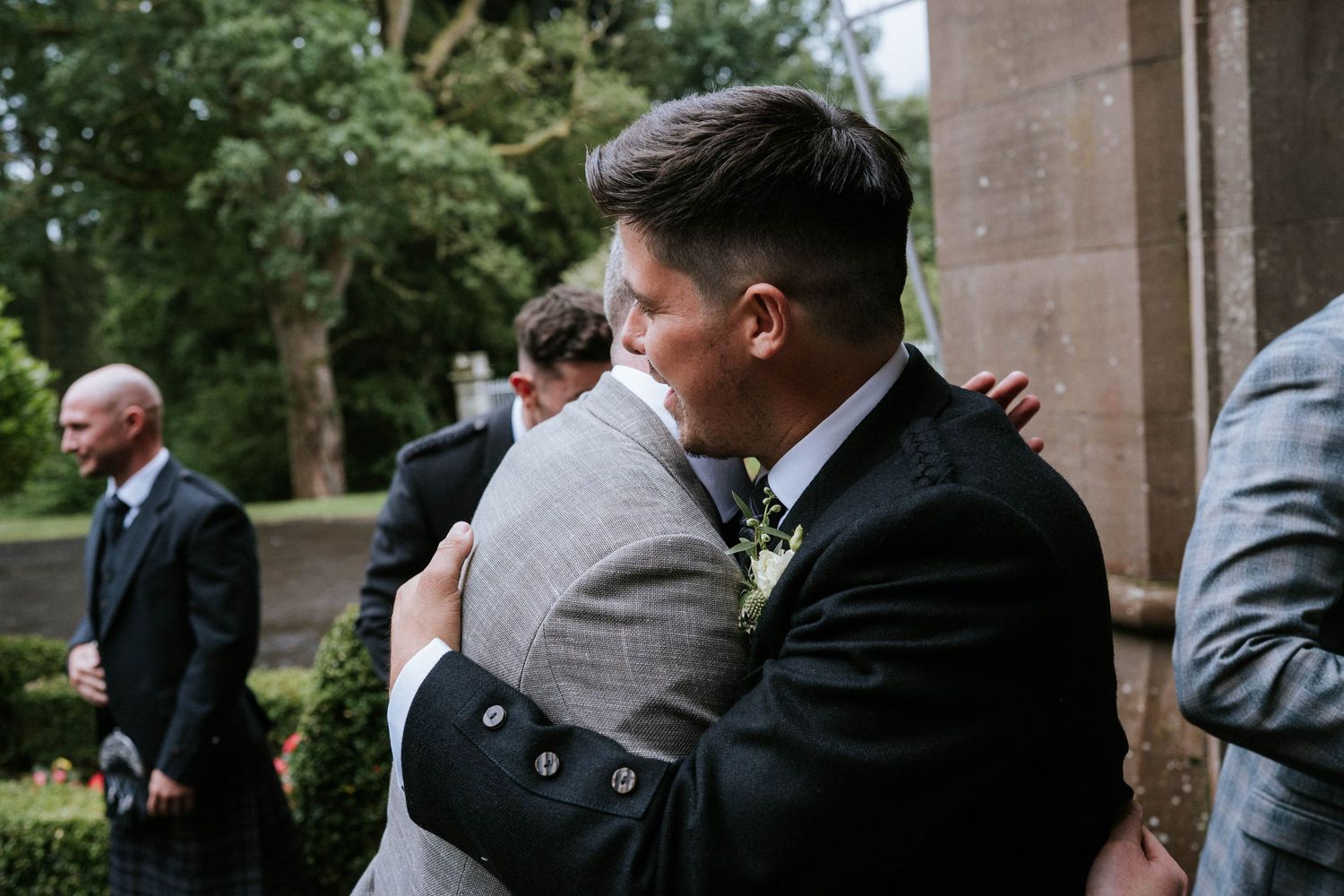 Wedding party members stand together outside a stone building in formal attire.