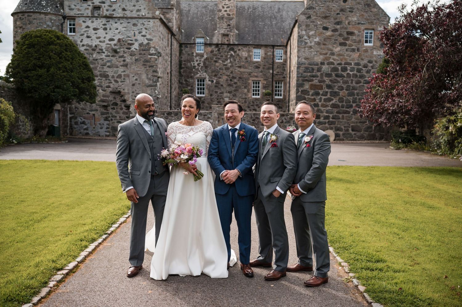 Wedding party poses in front of stone building with green lawn.