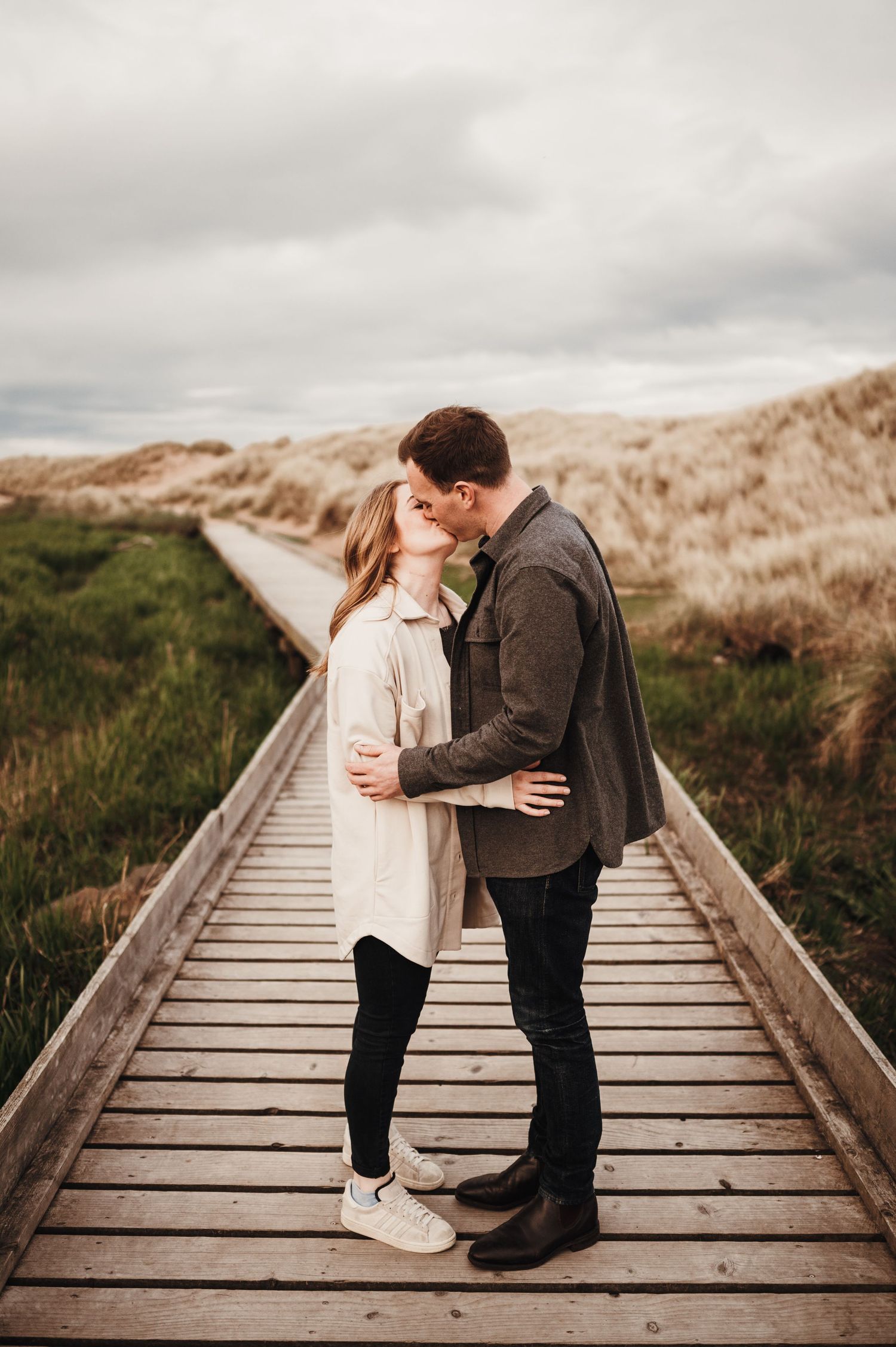 A couple shares a romantic moment on a wooden boardwalk path through coastal dunes on an overcast day.