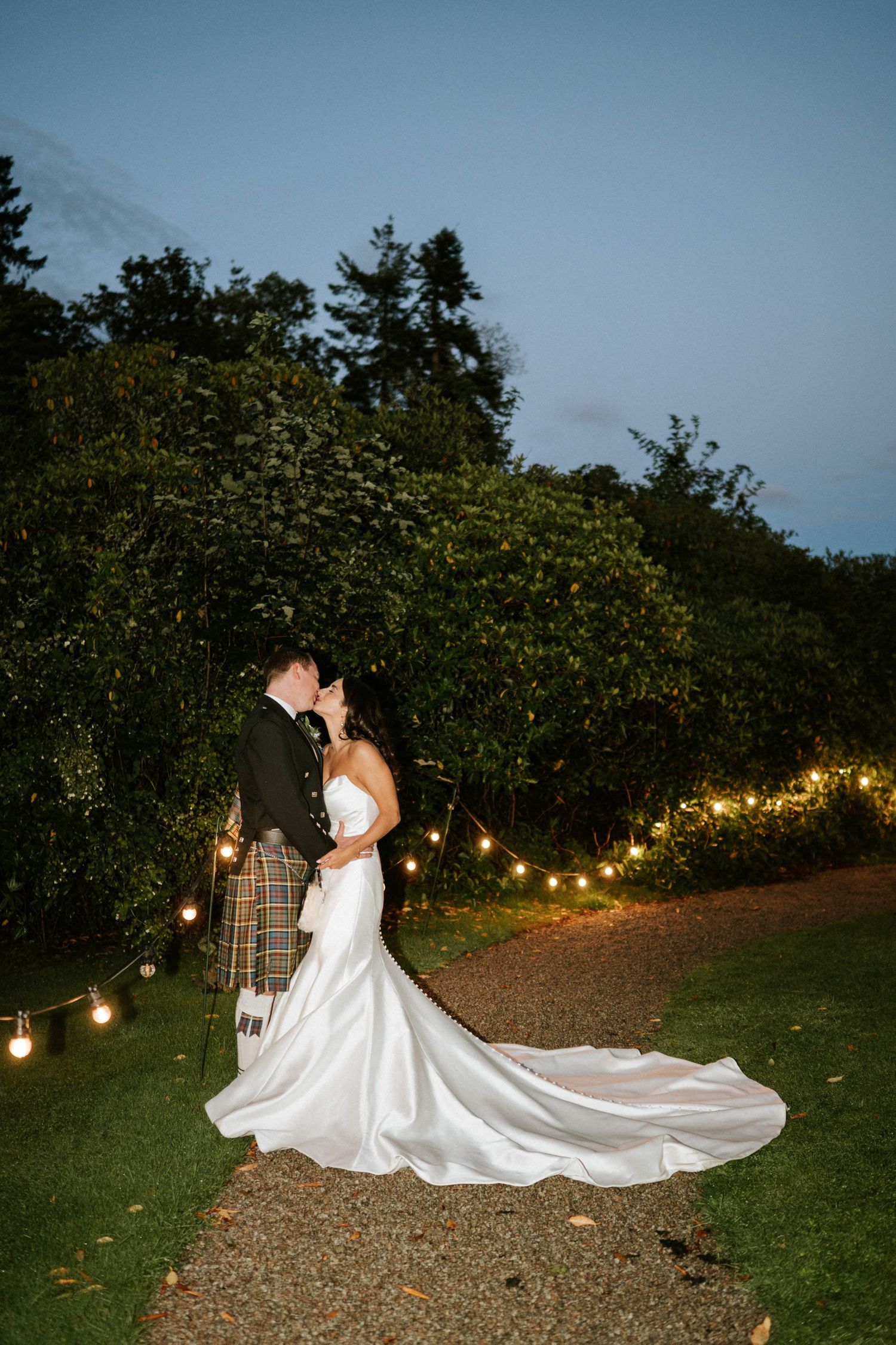 Evening outdoor wedding portrait with string lights and flowing white dress on garden path.