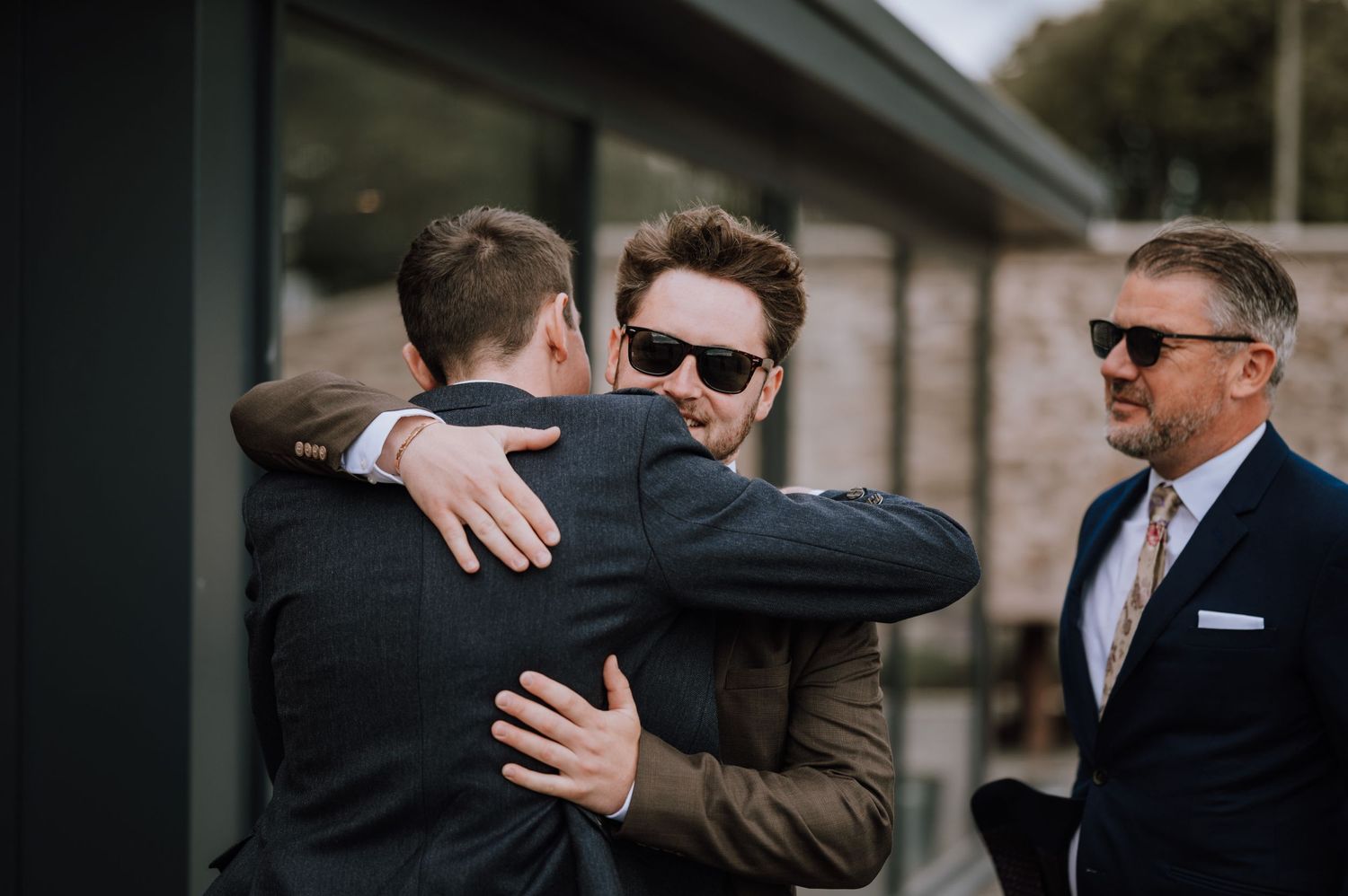 Well-dressed men share emotional embrace at outdoor wedding celebration.