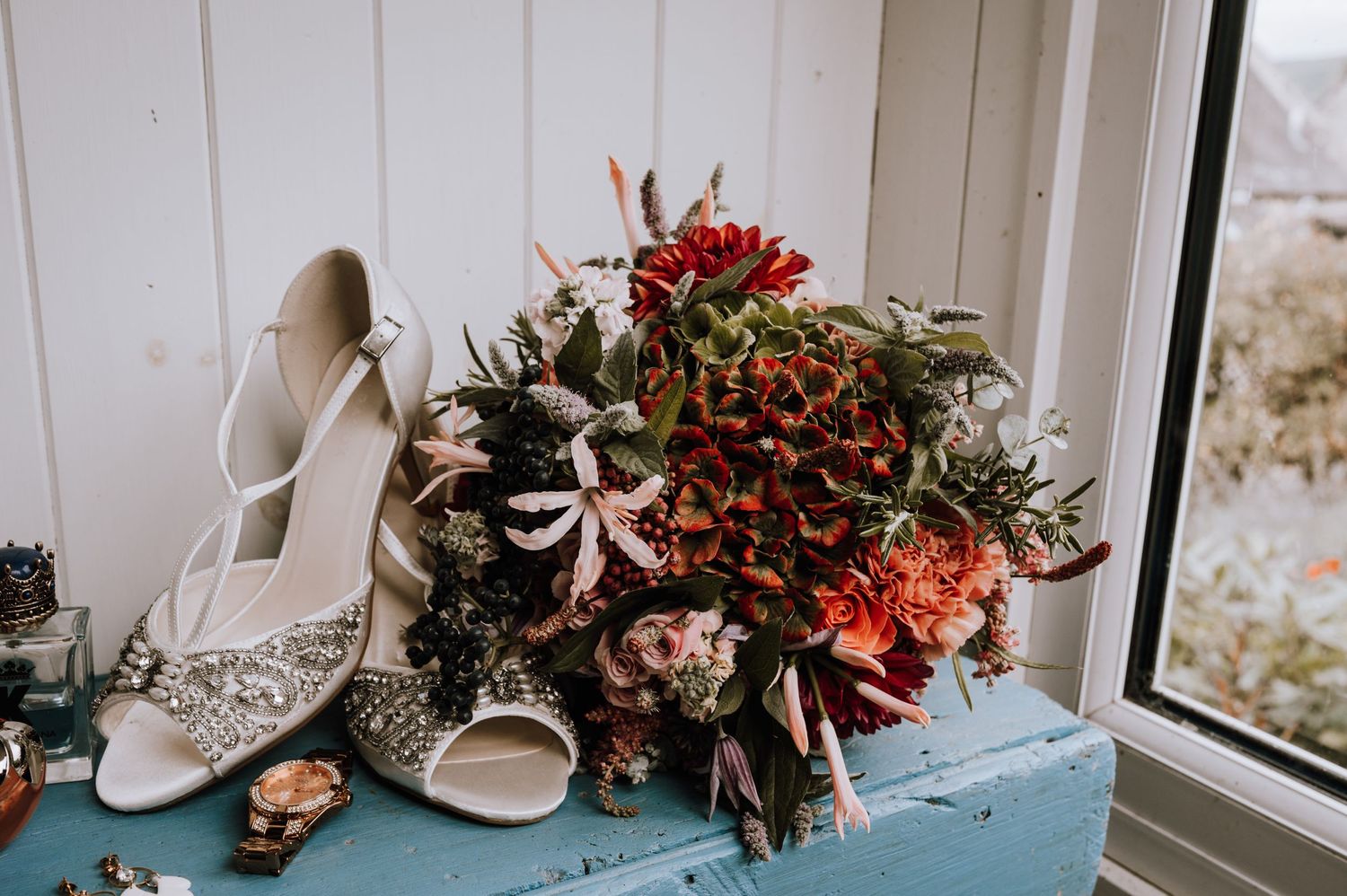 Wedding accessories including lace shoes and red floral bouquet on blue shelf near window.