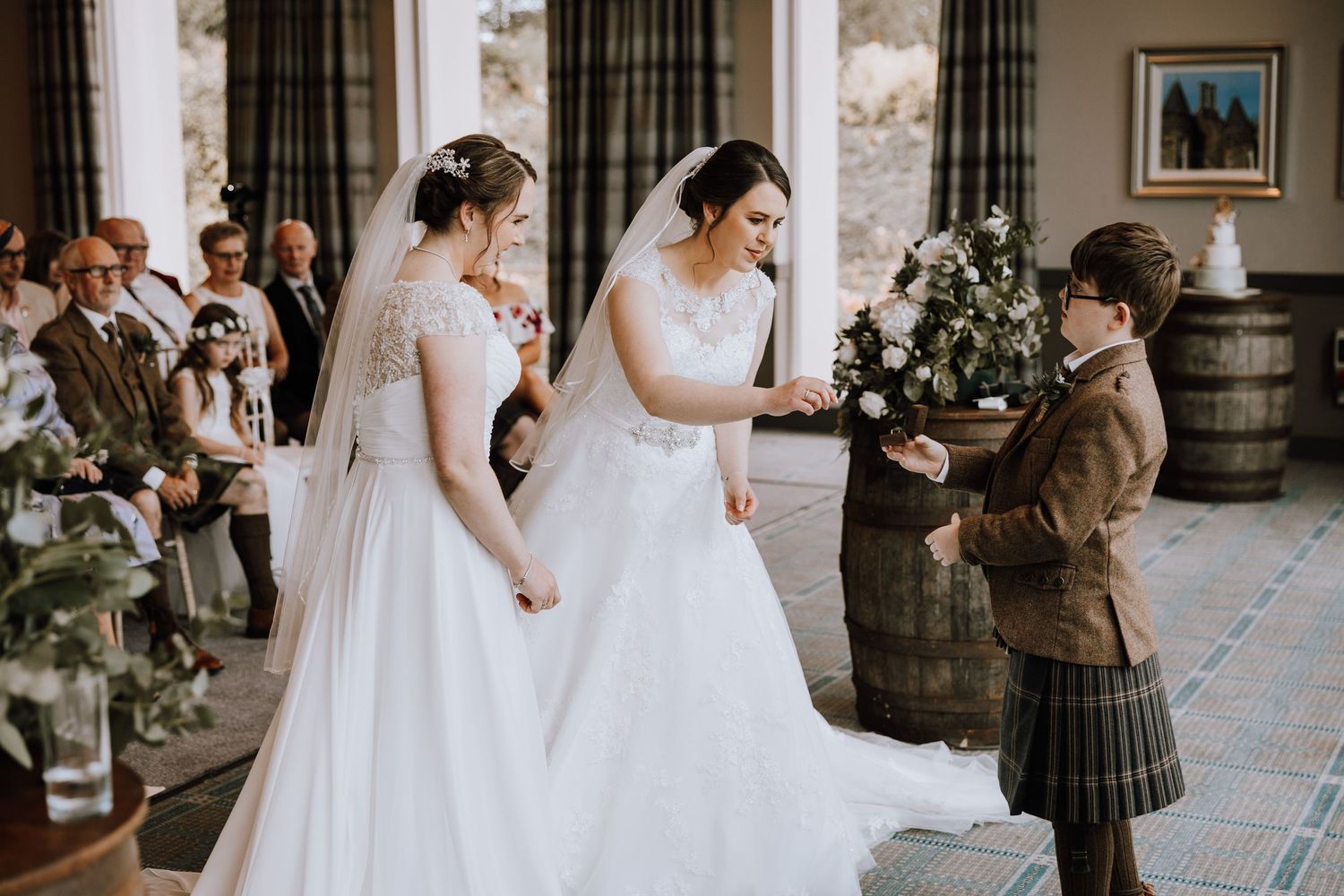 Two brides in white wedding dresses interact with a guest wearing a traditional Scottish kilt during a wedding ceremony.