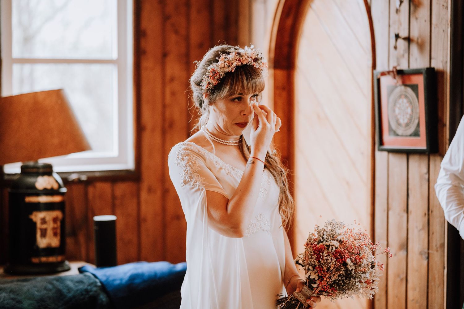 A bride in a white lace dress wipes away tears while holding a pink bouquet.