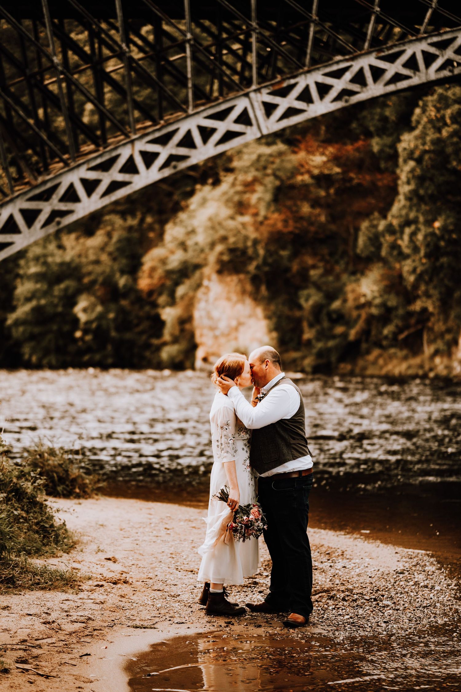 A romantic wedding couple embraces by a river beneath a white metal bridge in a moody autumn setting.