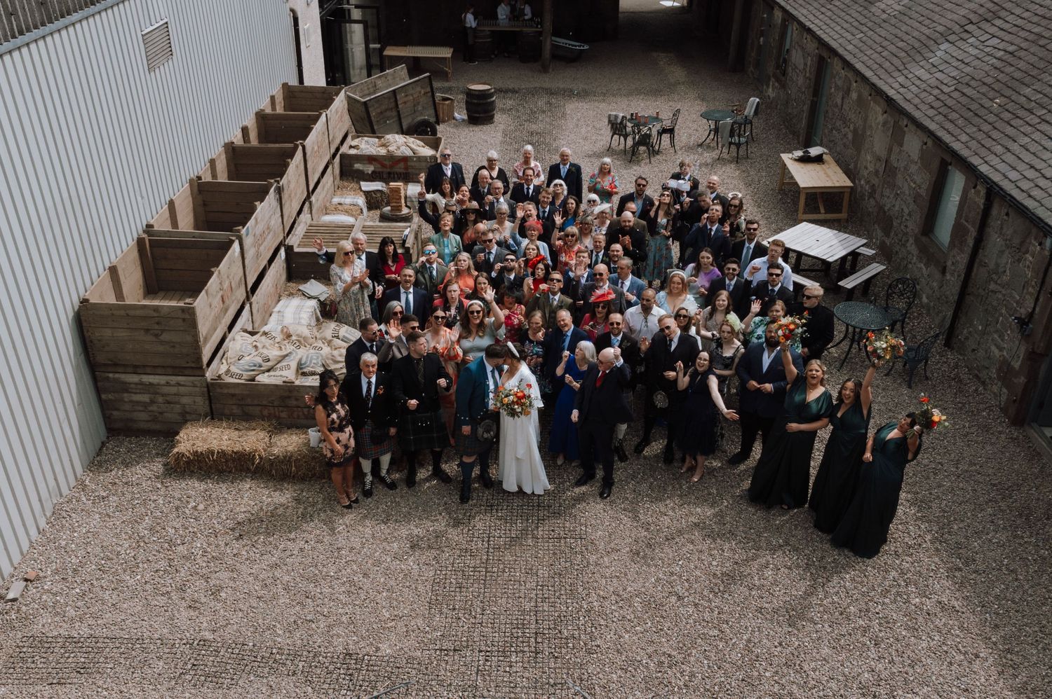 Aerial view of wedding party and guests gathered for a group photo outside a rustic venue.