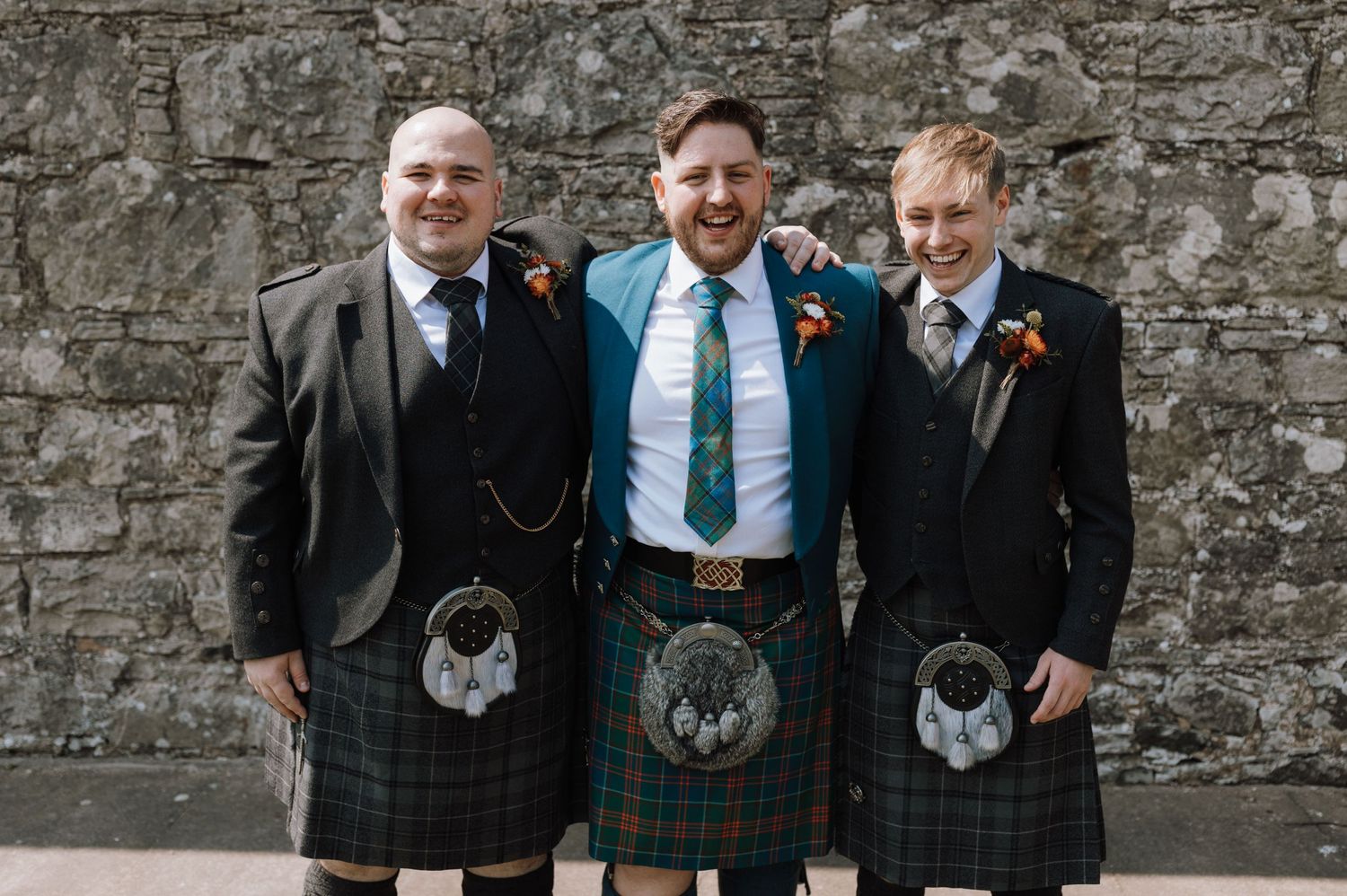 Three people in kilts and formal attire smiling for wedding photo.