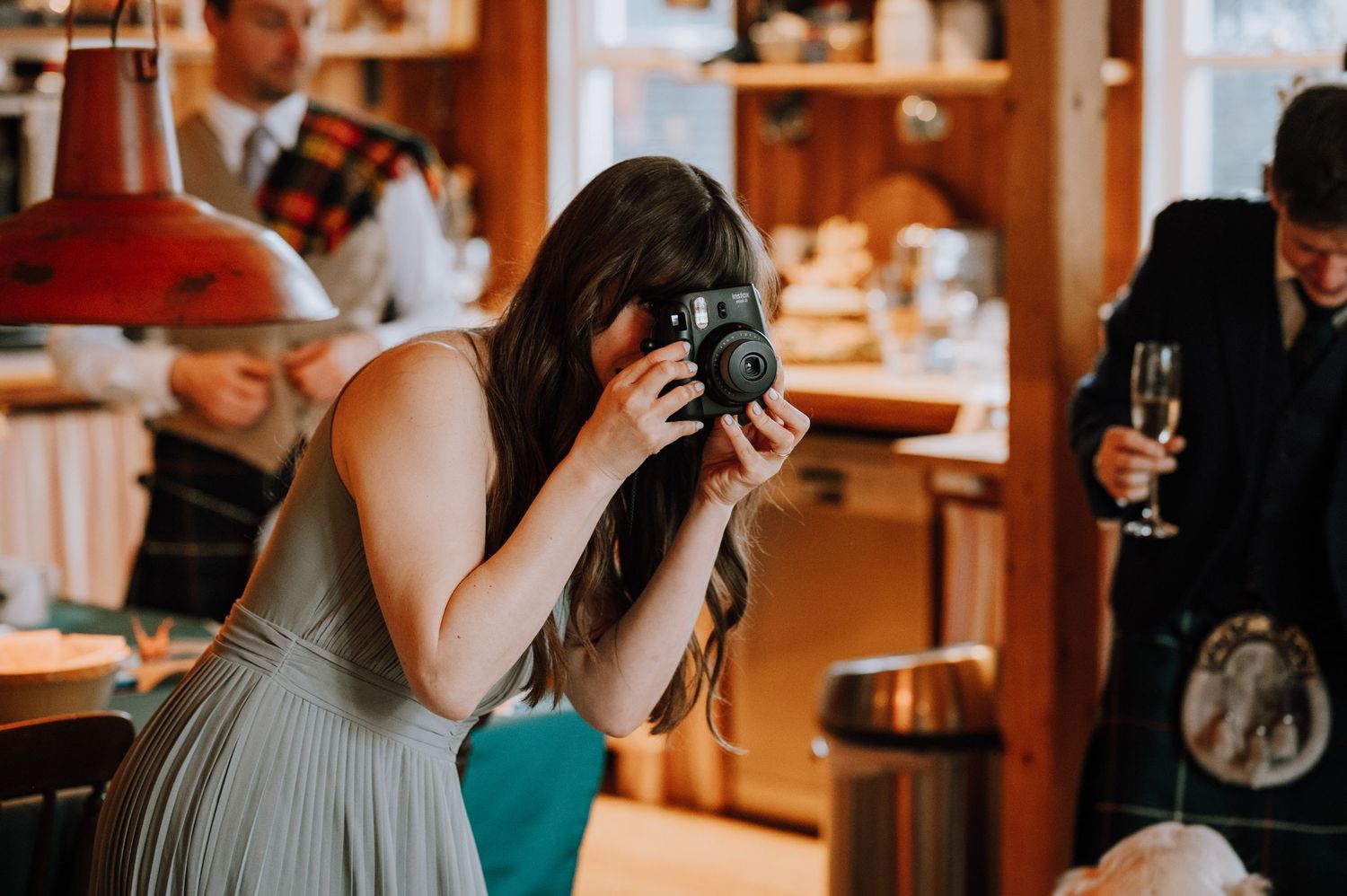 Guest in grey dress takes photos during wedding reception in wooden interior.