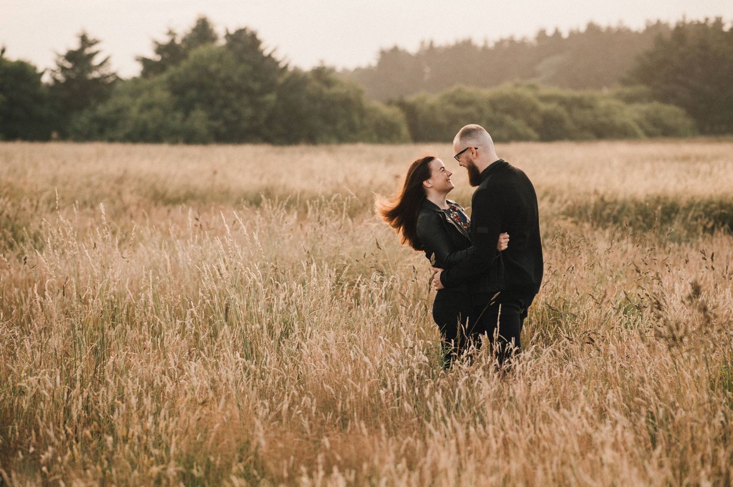 A romantic moment captured in a golden field of tall grass at sunset with soft lighting and pine trees in background.