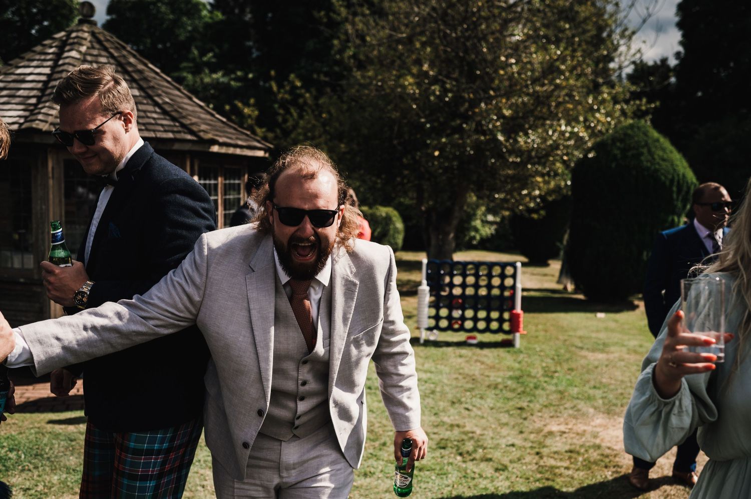 Wedding guests mingling outdoors on a sunny day with one wearing a light colored suit and sunglasses.