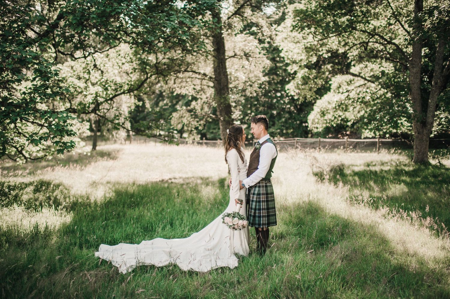 A romantic wedding couple poses in a sunlit forest meadow with the groom wearing traditional Scottish tartan kilt attire.