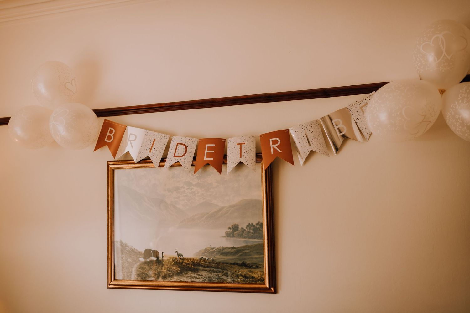 Gold 'BRIDE TRIBE' banner hanging on wall above framed landscape painting with white balloons.