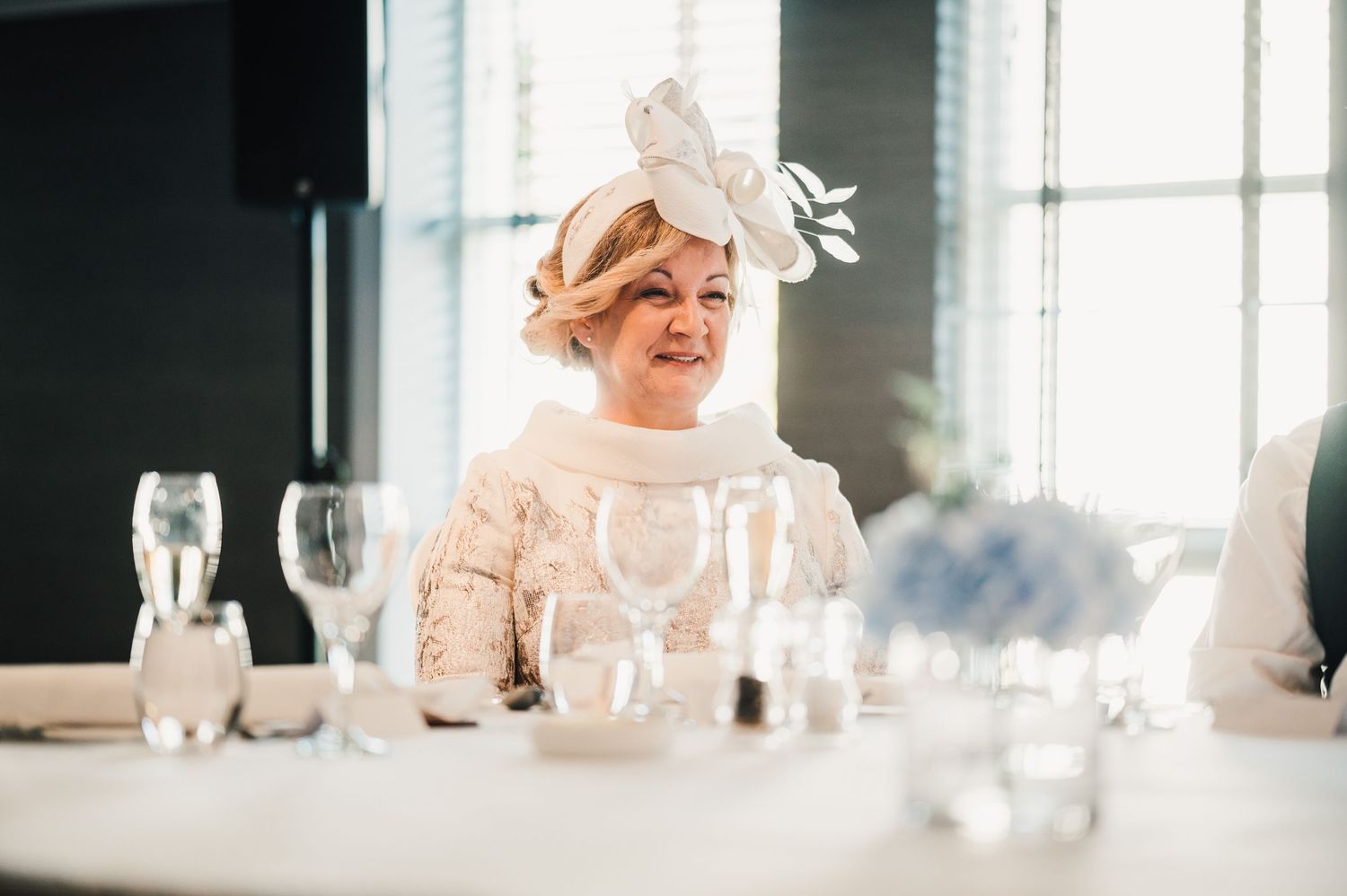 An elegant wedding guest in light attire smiles while seated at a reception table with wine glasses.