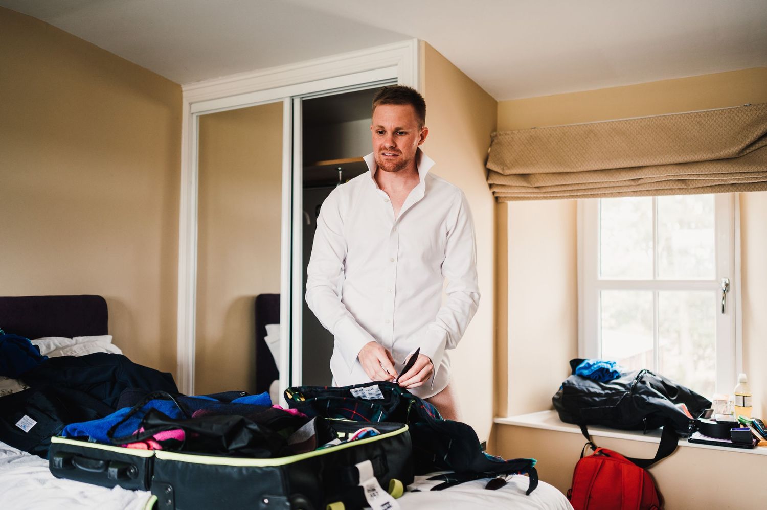A person in a white shirt smiles while getting ready in a bedroom with luggage and clothes visible.