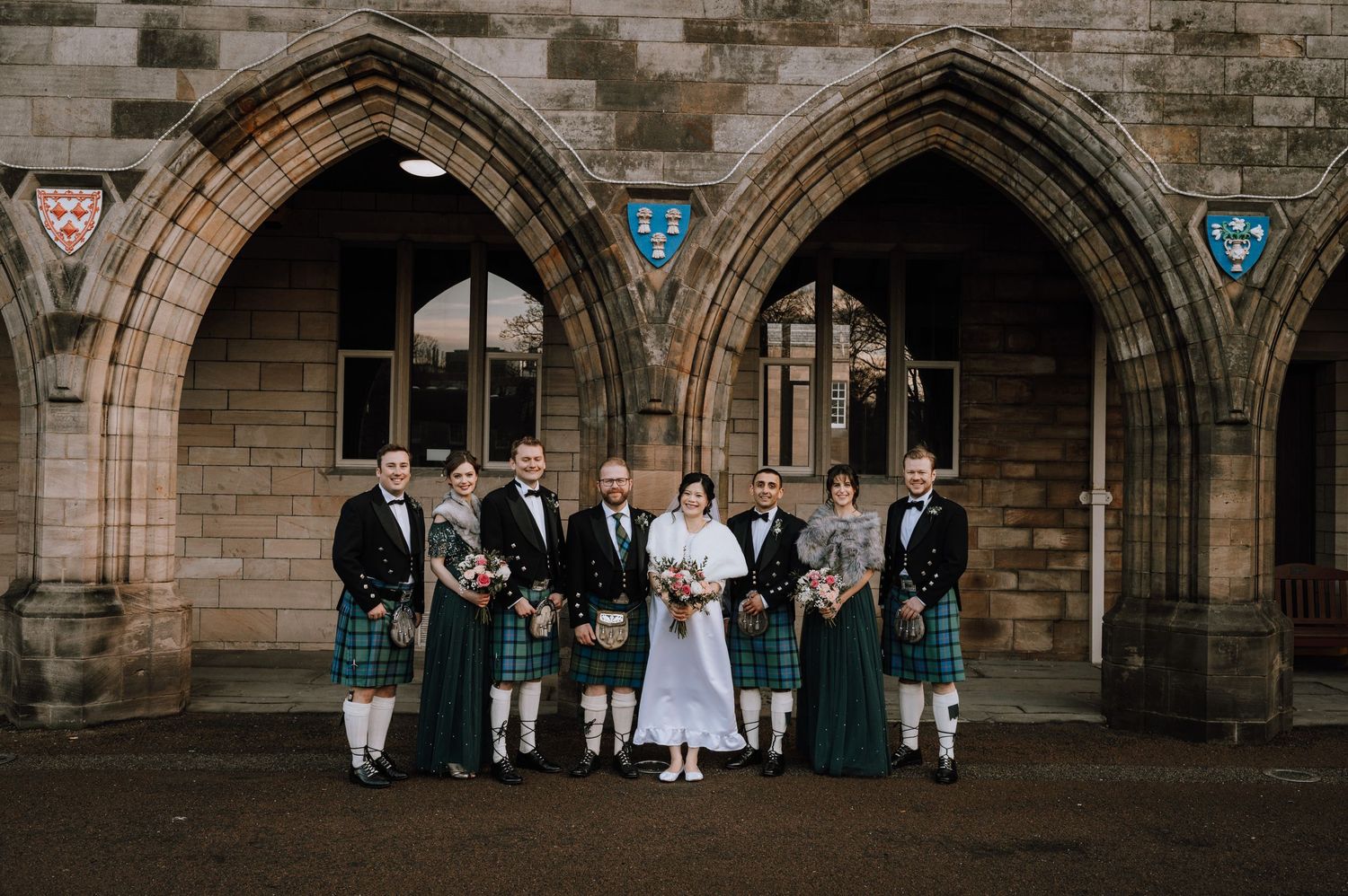 A bridal party with men in kilts stands beneath Gothic stone arches of a church.