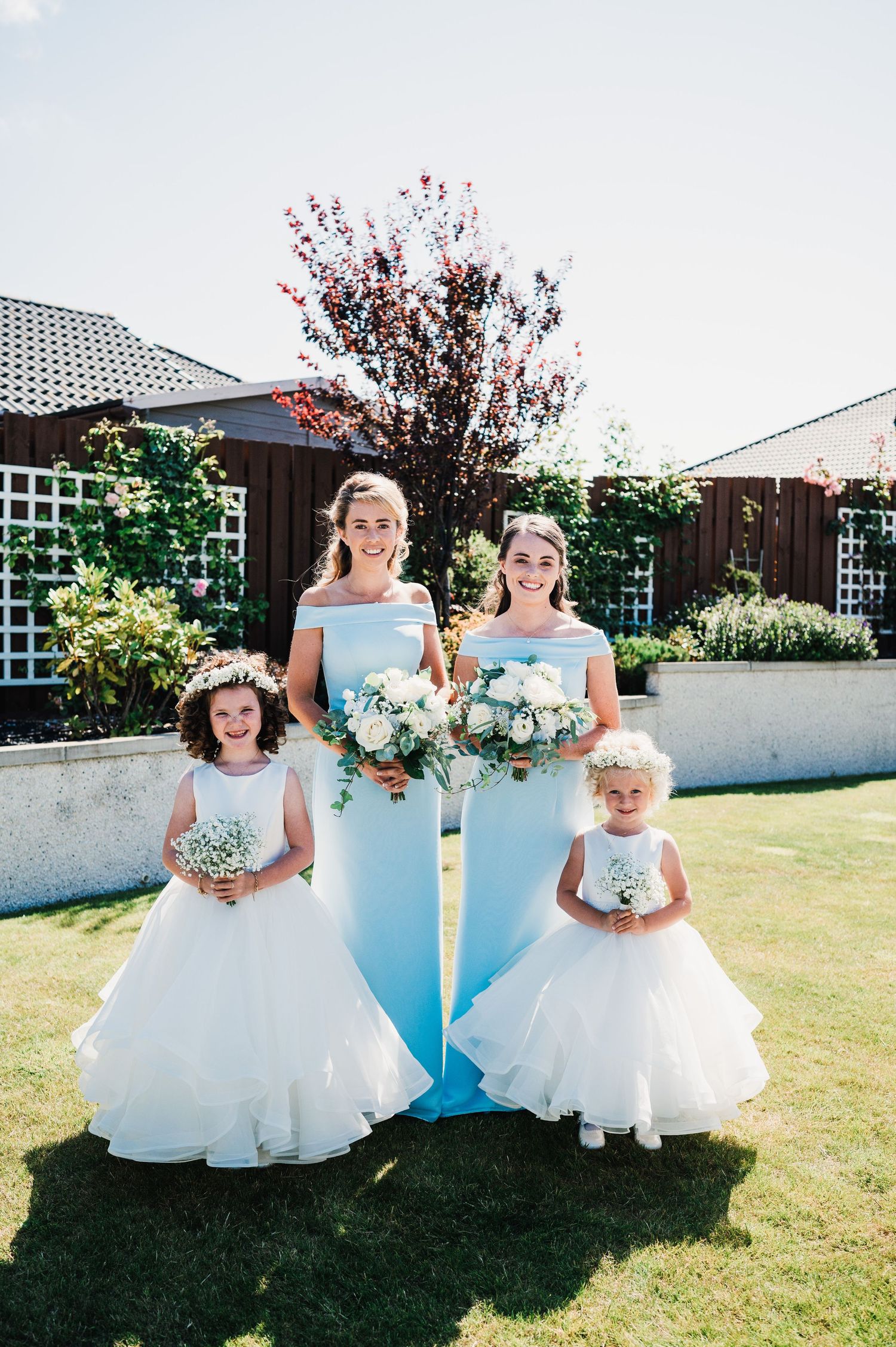 Wedding party members in light blue dresses and white flower girl dresses pose together in a garden setting.