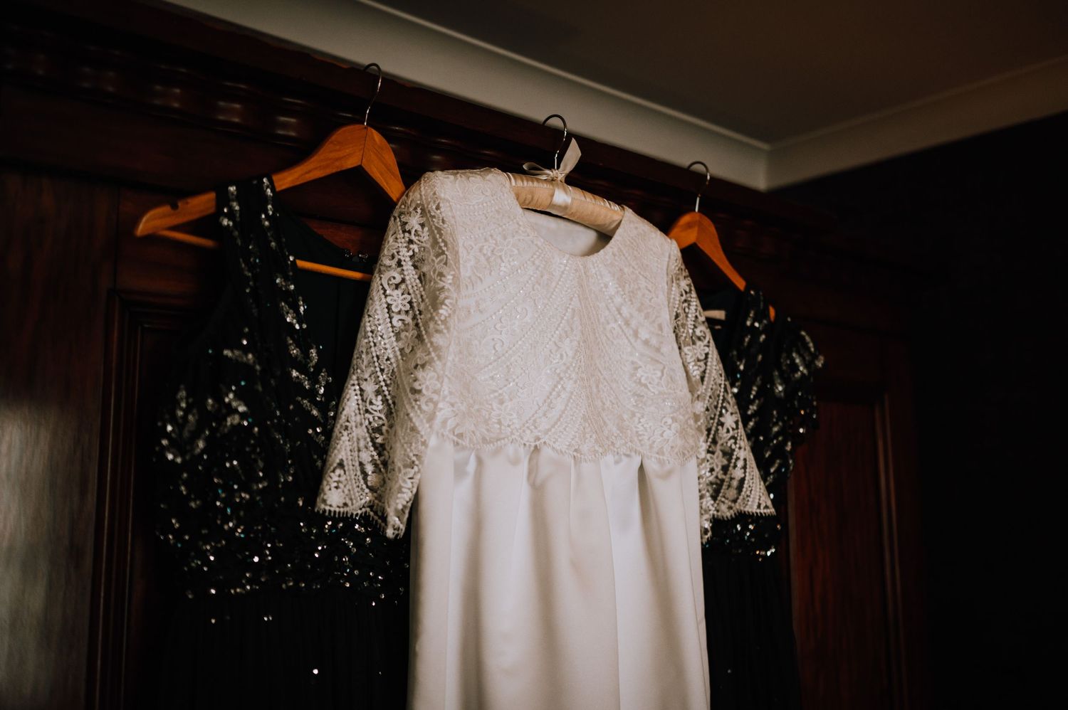 White lace wedding dress hanging on orange hangers against dark wooden wardrobe.