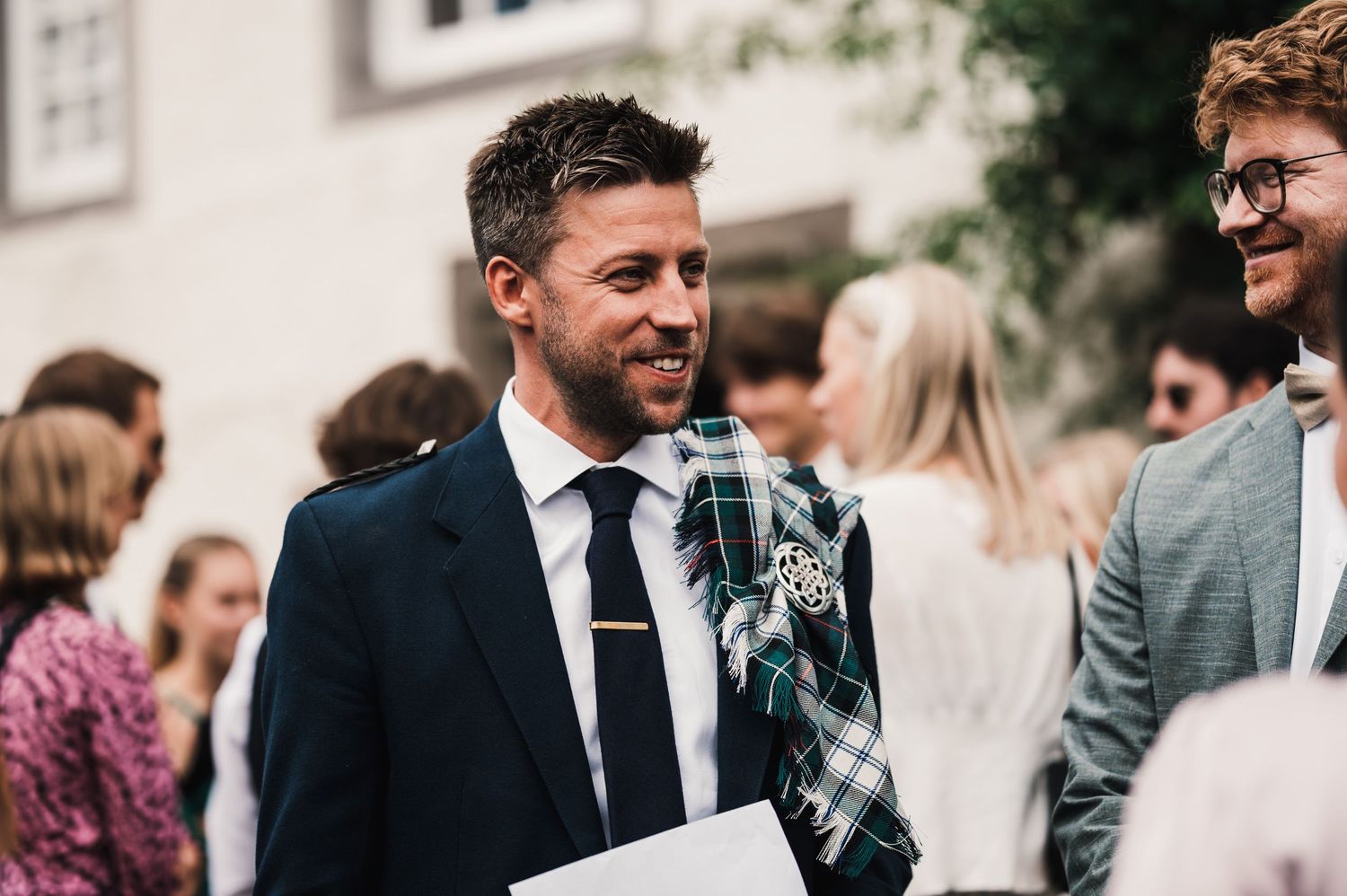 Wedding guests mingle at an outdoor reception with person wearing navy suit and tie in foreground.