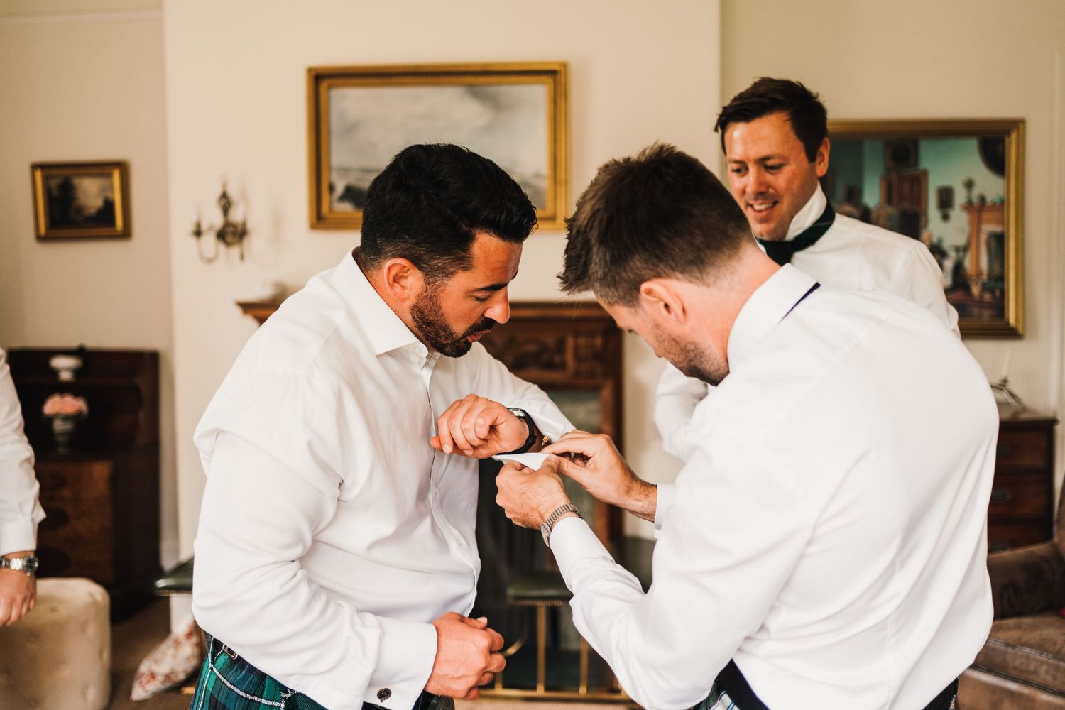 Groomsmen helping each other prepare in a formal room with white shirts.