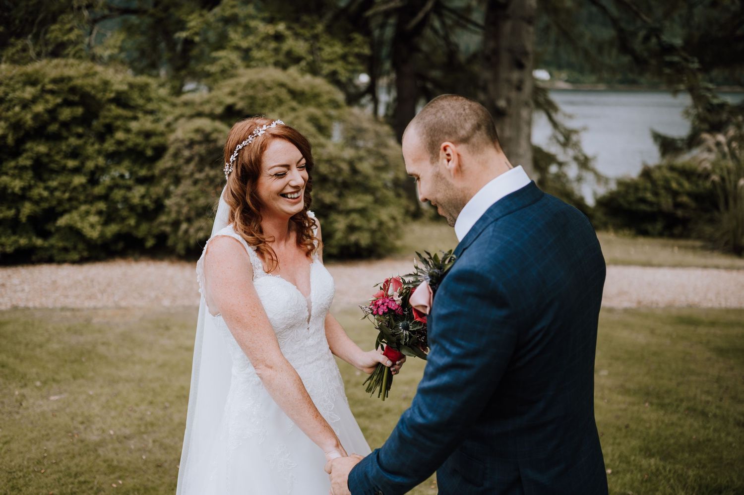 Bride and groom laugh together outdoors with bouquet and greenery.