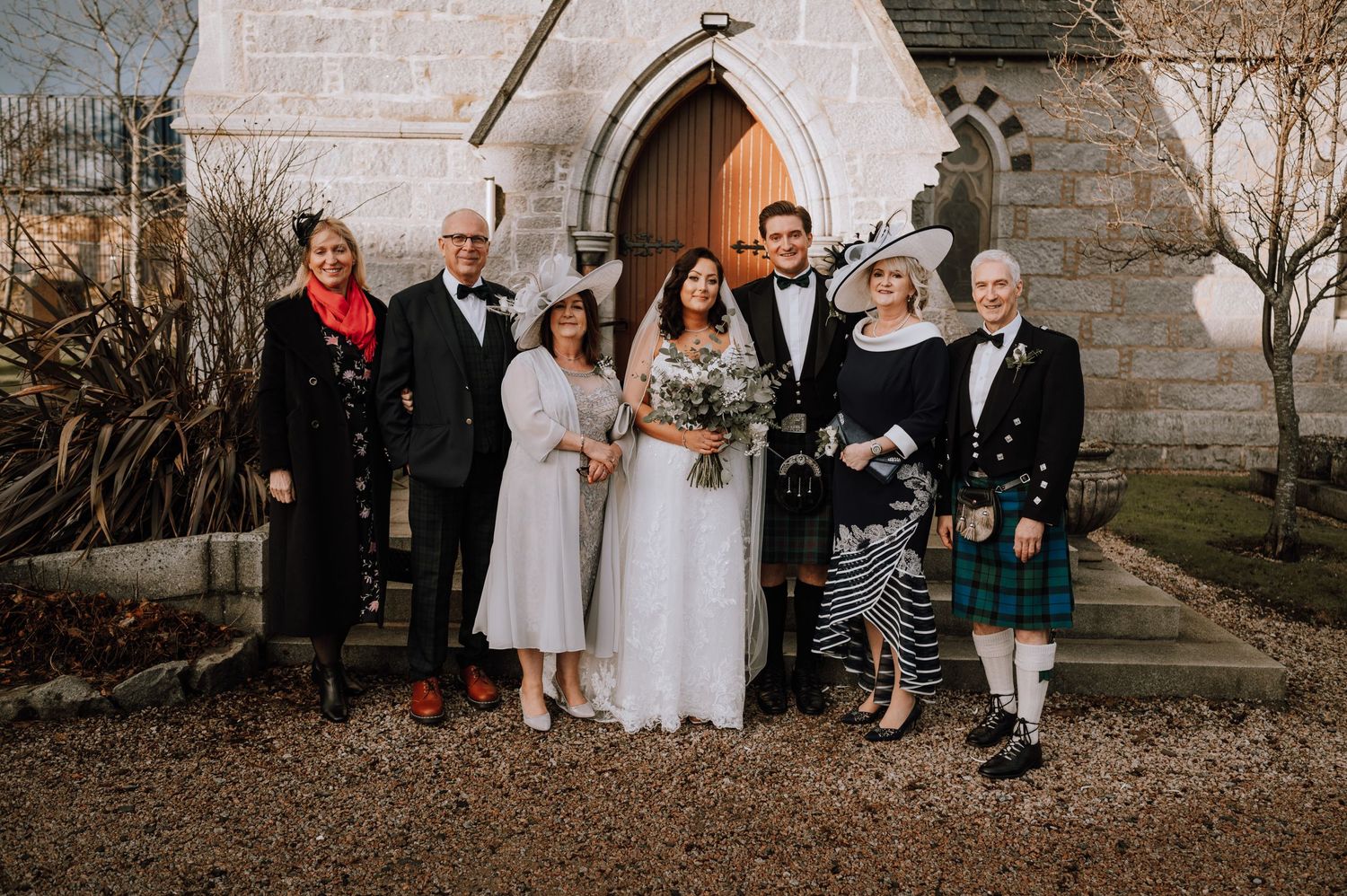 Wedding party poses outside stone church entrance with Scottish kilts and formal attire.