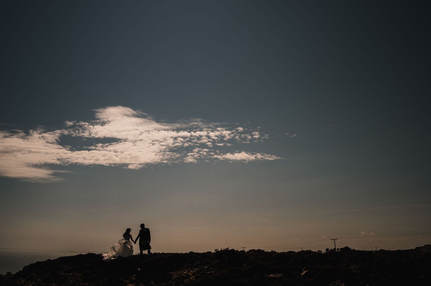 Silhouetted couple stands together against sunset sky on hilltop.