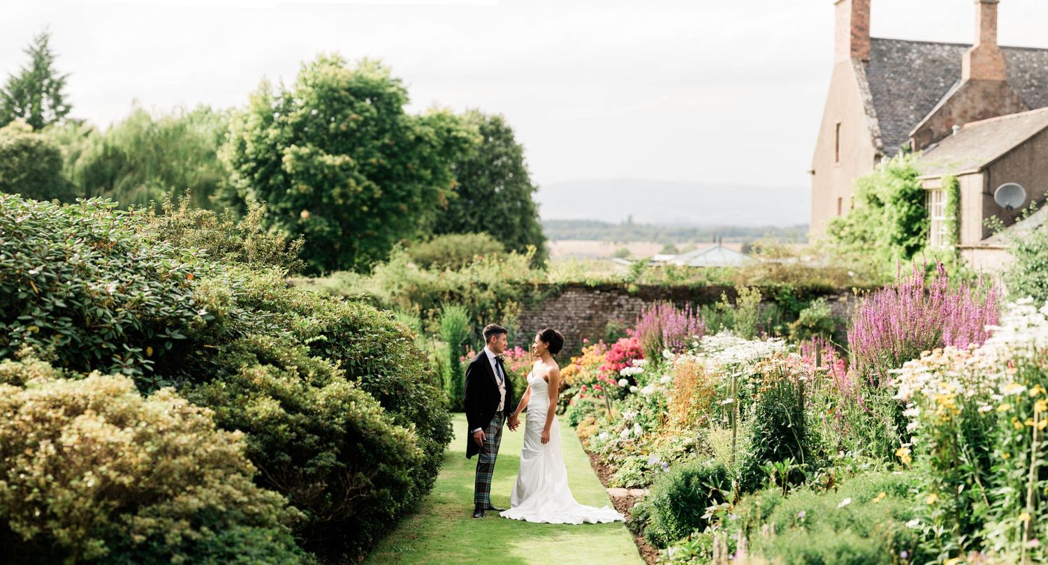 A wedding couple walks through a stunning garden path with stone house in background.