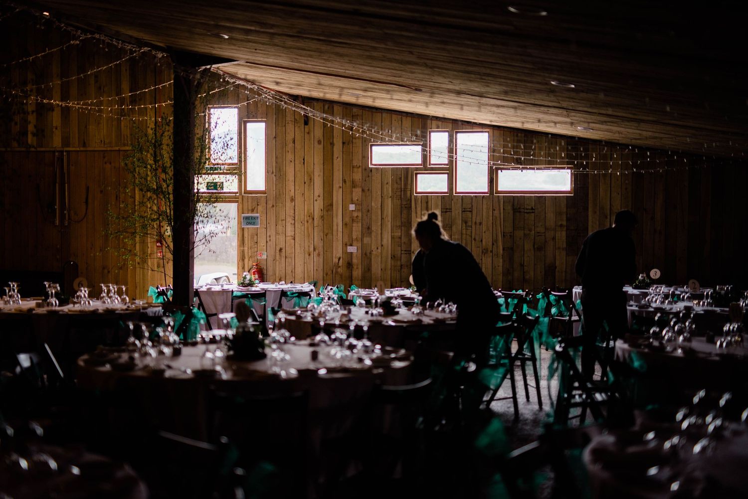 Dark reception hall with tables set for wedding dinner under dramatic window lighting.